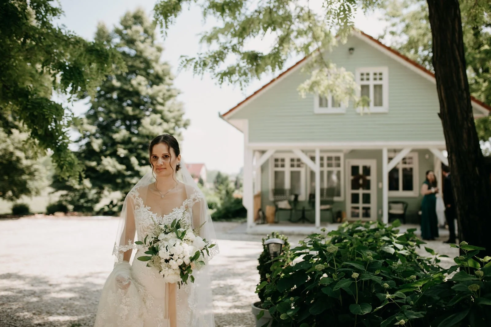 A bride in a white wedding gown holding a bouquet of white flowers stands outside in front of a green house with trees around on a bright day.