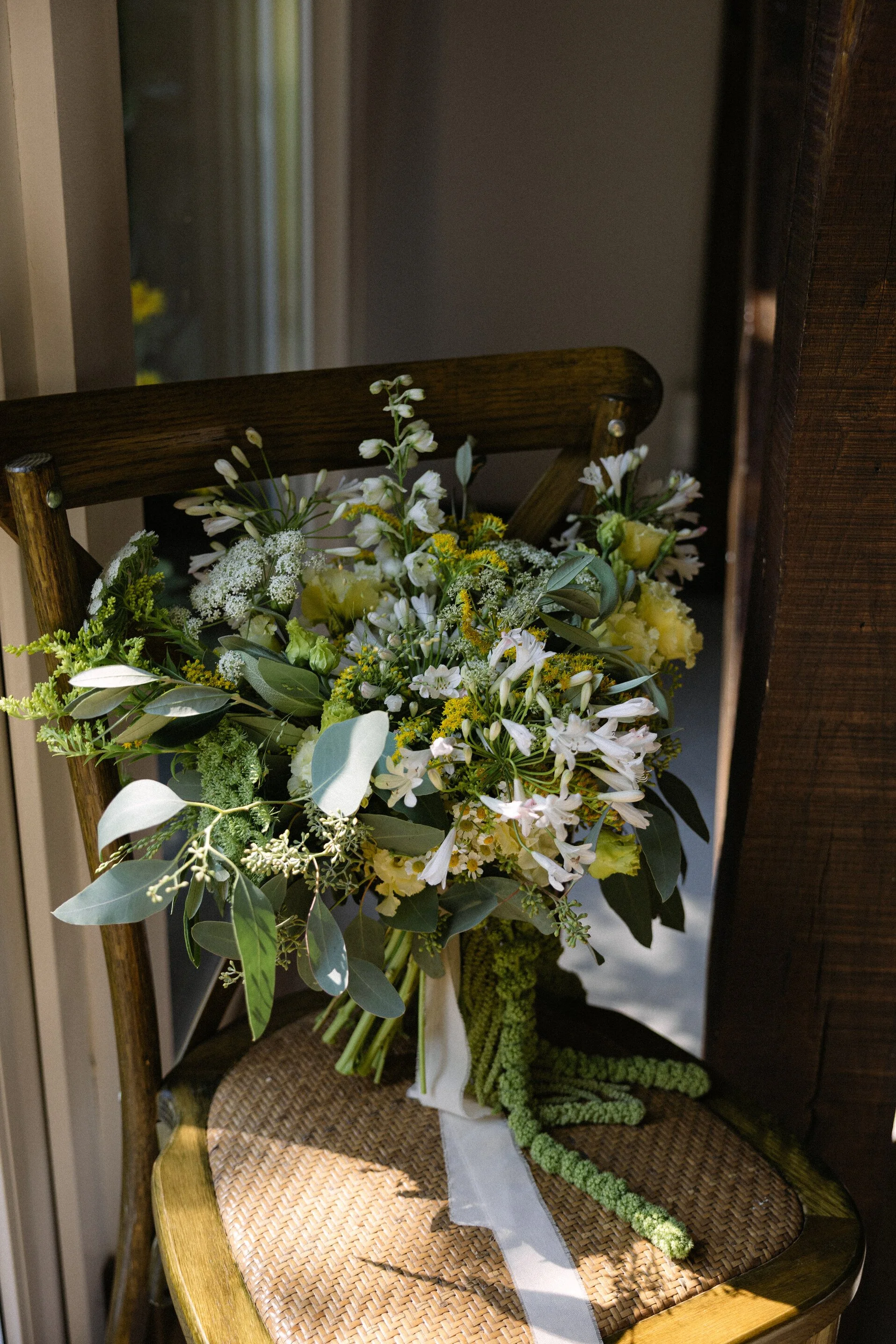 A bouquet of white and yellow flowers with green leaves resting on a wooden chair with a woven seat, near a window.