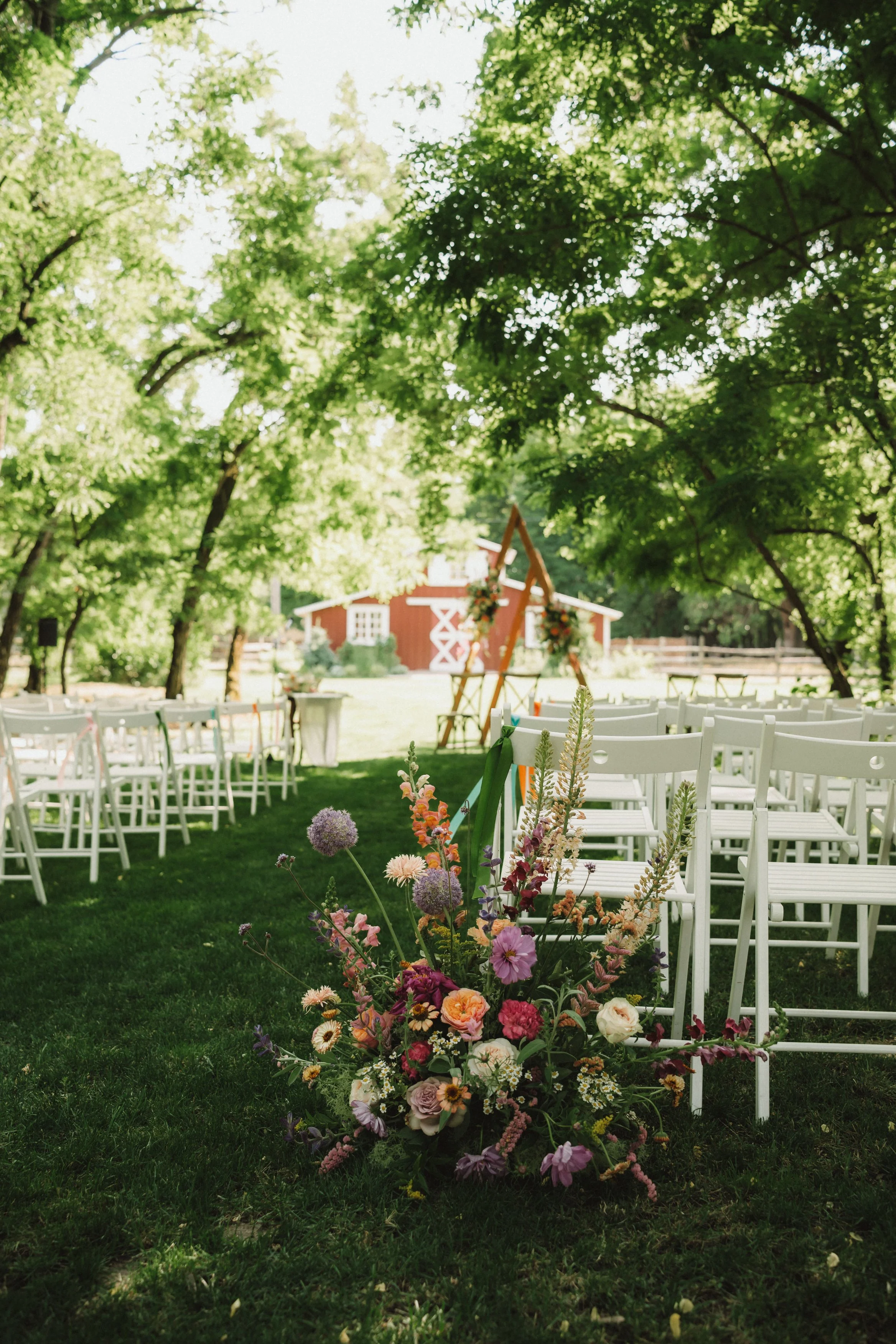 Szabadtéri esküvői helyszín fehér székekkel, rendezve a fűben, zöld fákkal körülvéve. Az előtérben színes virágkompozíció látható, a háttérben vörös pajta épület.
