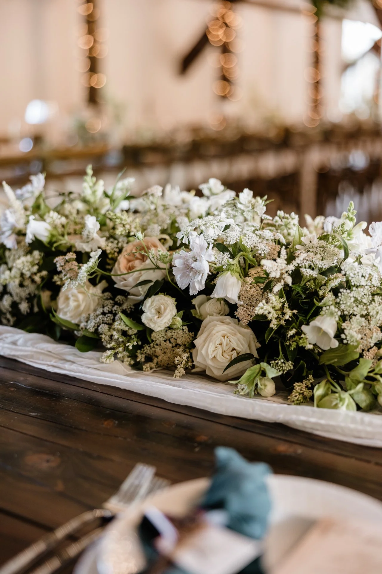 Elegáns virágkompozíció fehér rózsákkal, gyöngyvirággal és zöld növényzettel egy fa asztalon, díszített rendezvényhelyszínen.
