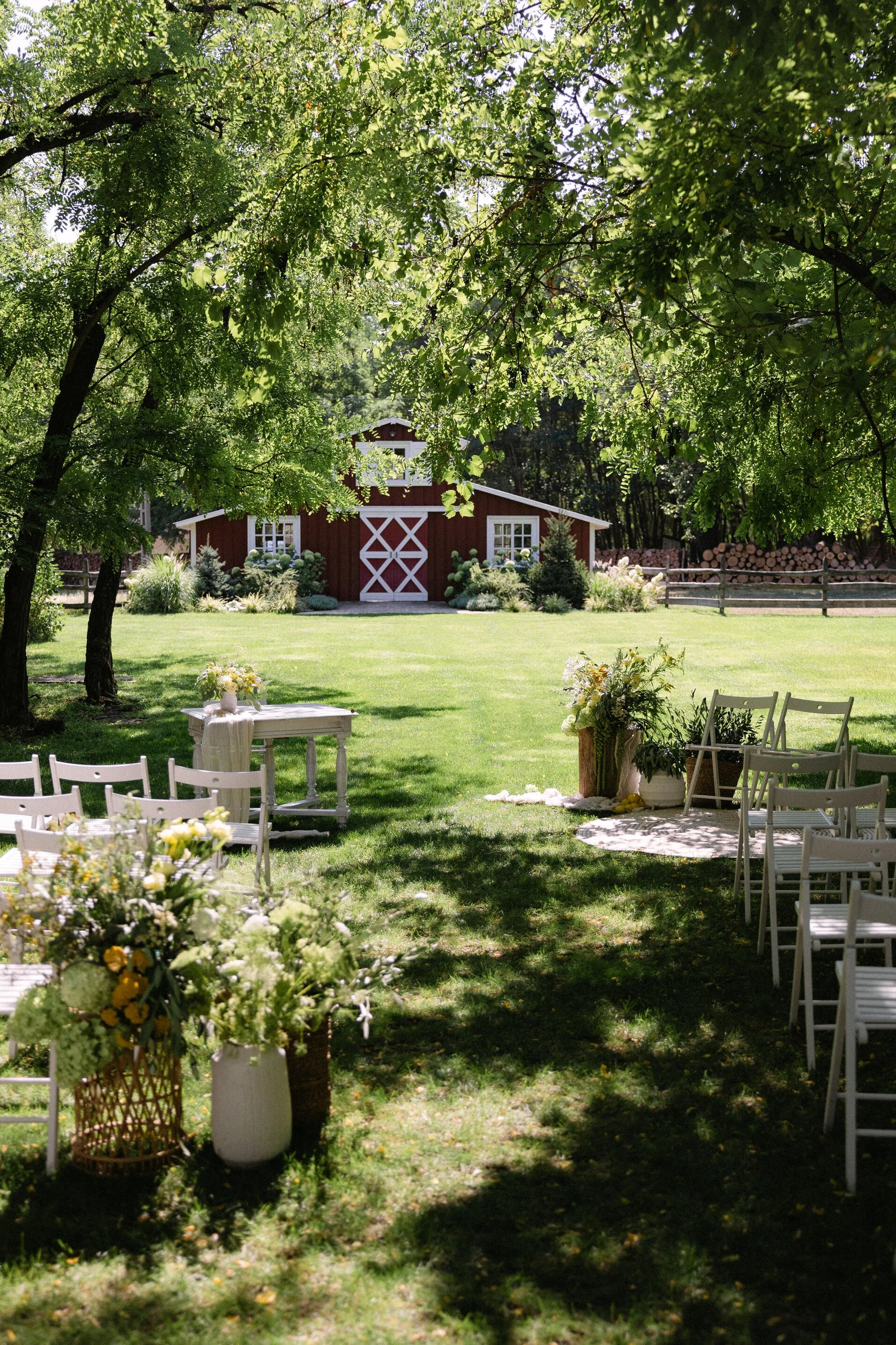 Outdoor wedding setup with white chairs and floral arrangements, leading to a red barn in a green, sunny garden.