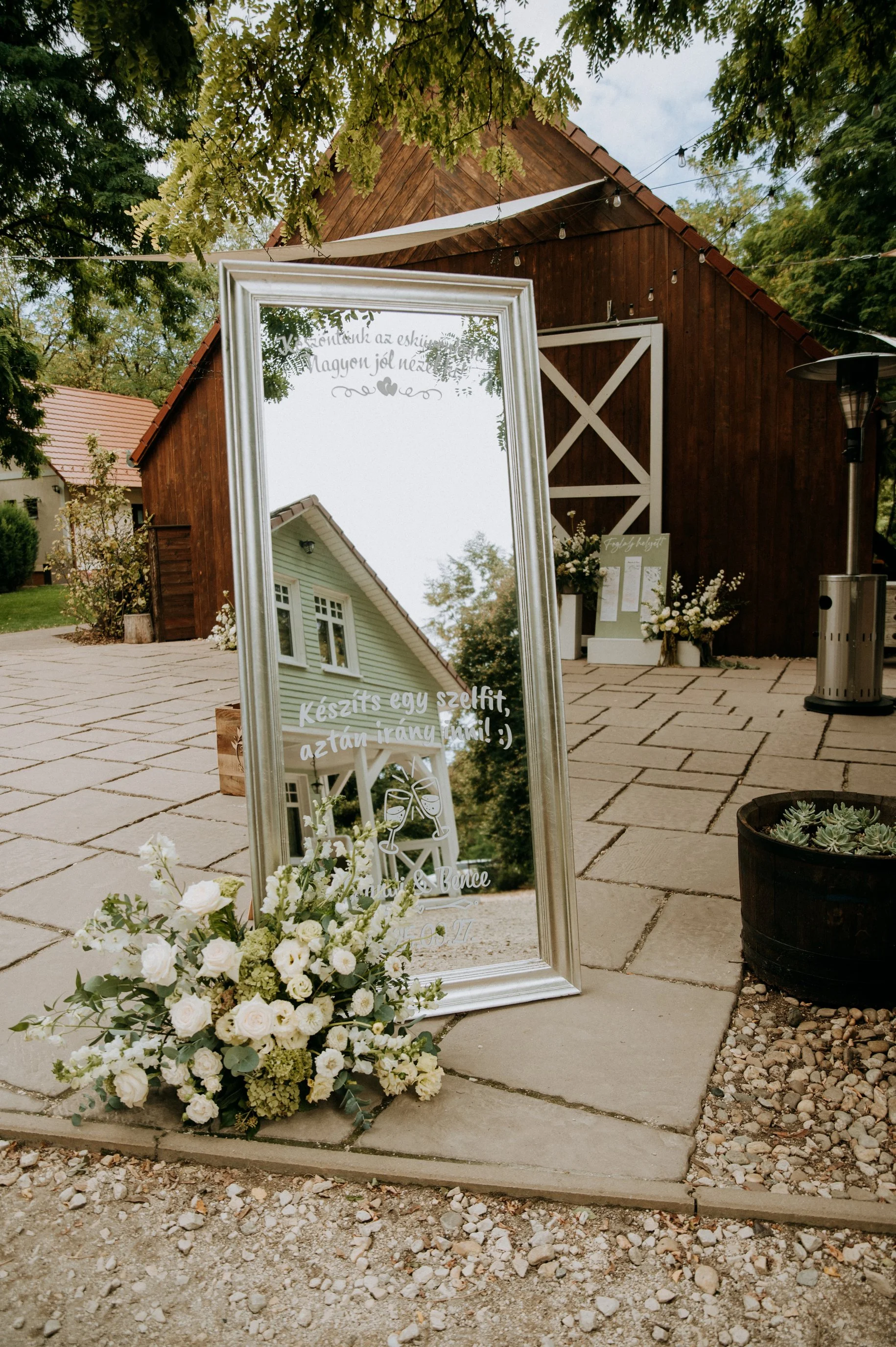 A large standing mirror with handwritten Hungarian text, decorated with white flowers at the base, outdoors on a cobblestone surface near a rustic wooden building with white door and window frames, surrounded by trees and string lights.