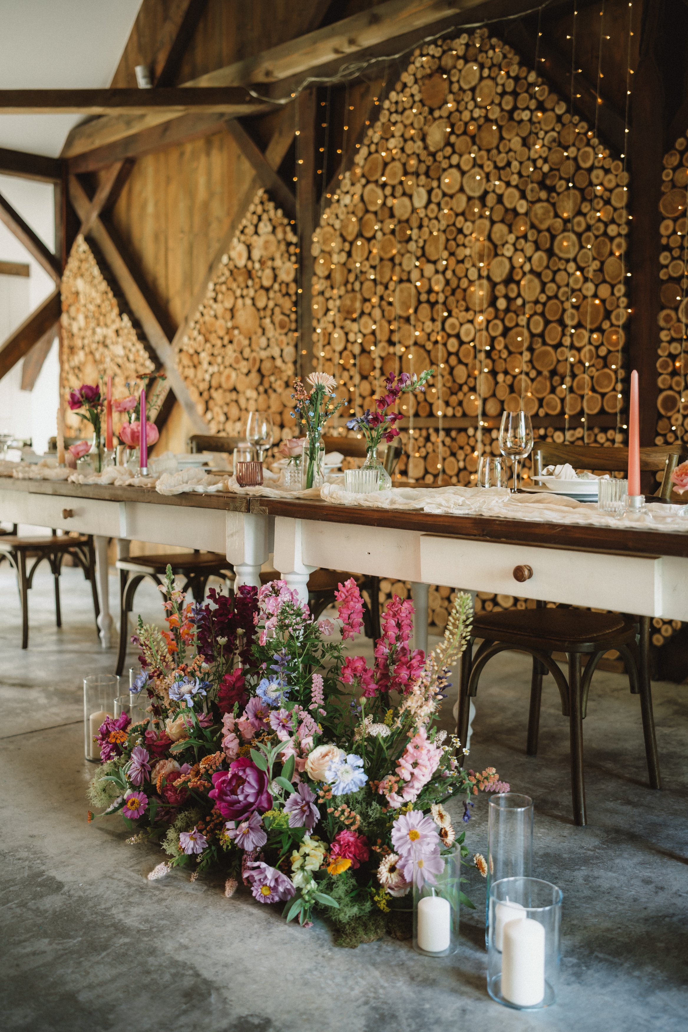 Rustic banquet table decorated with pink and purple flowers, candles, and place settings, set against a backdrop of stacked firewood and string lights.