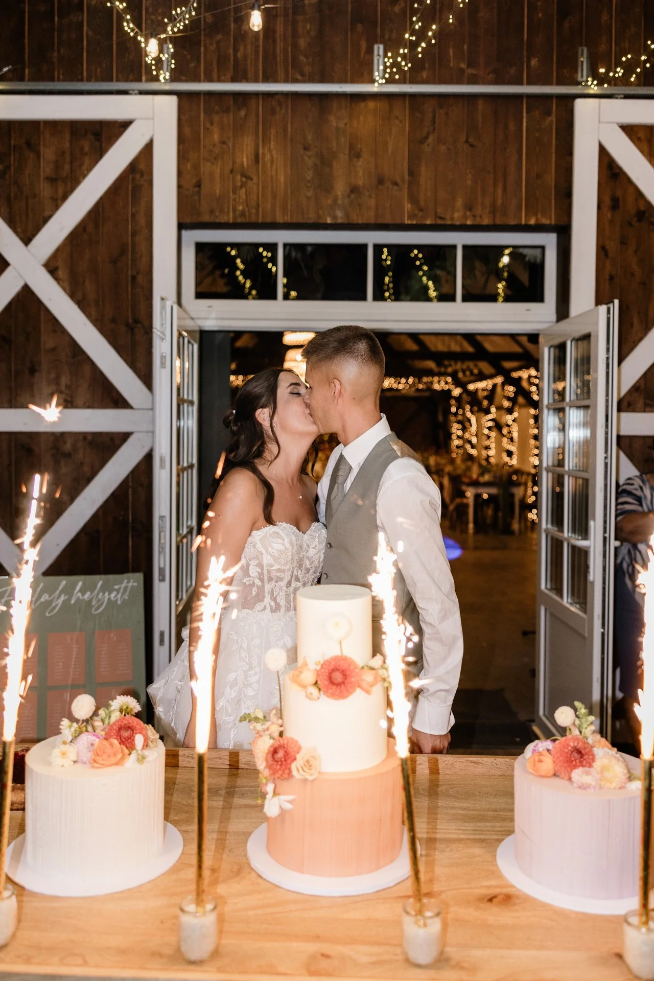 A menyasszony és a vőlegény csókolóznak az esküvői fogadásukon, miközben előttük egy rusztikus faasztalon három gyönyörűen díszített esküvői torta áll. A torták mellett csillagszórók égnek, amelyek látványos, ünnepi hangulatot teremtenek. 