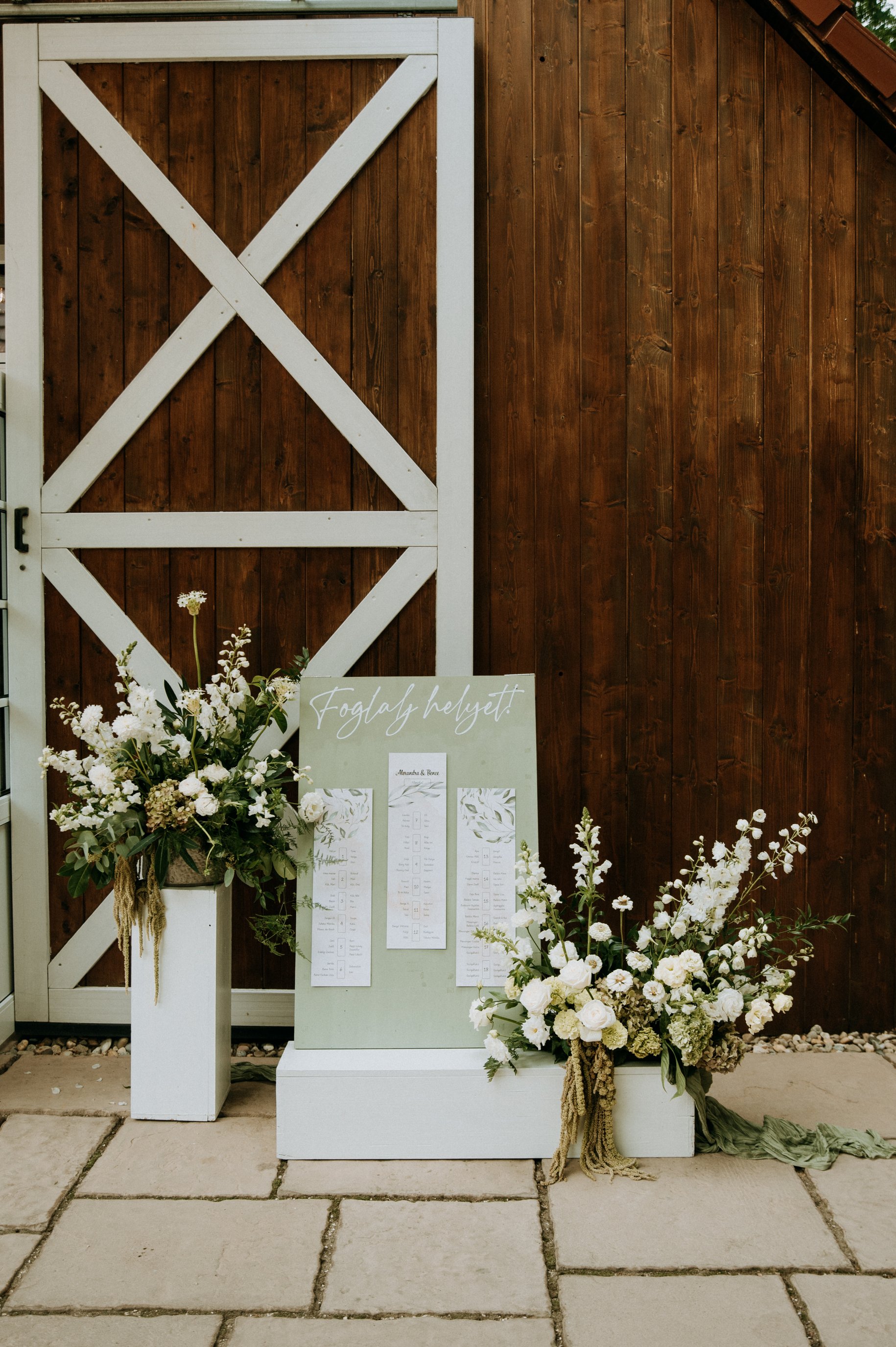 Decorative wedding seating chart display with white floral arrangements against a wooden wall and a white gate.
