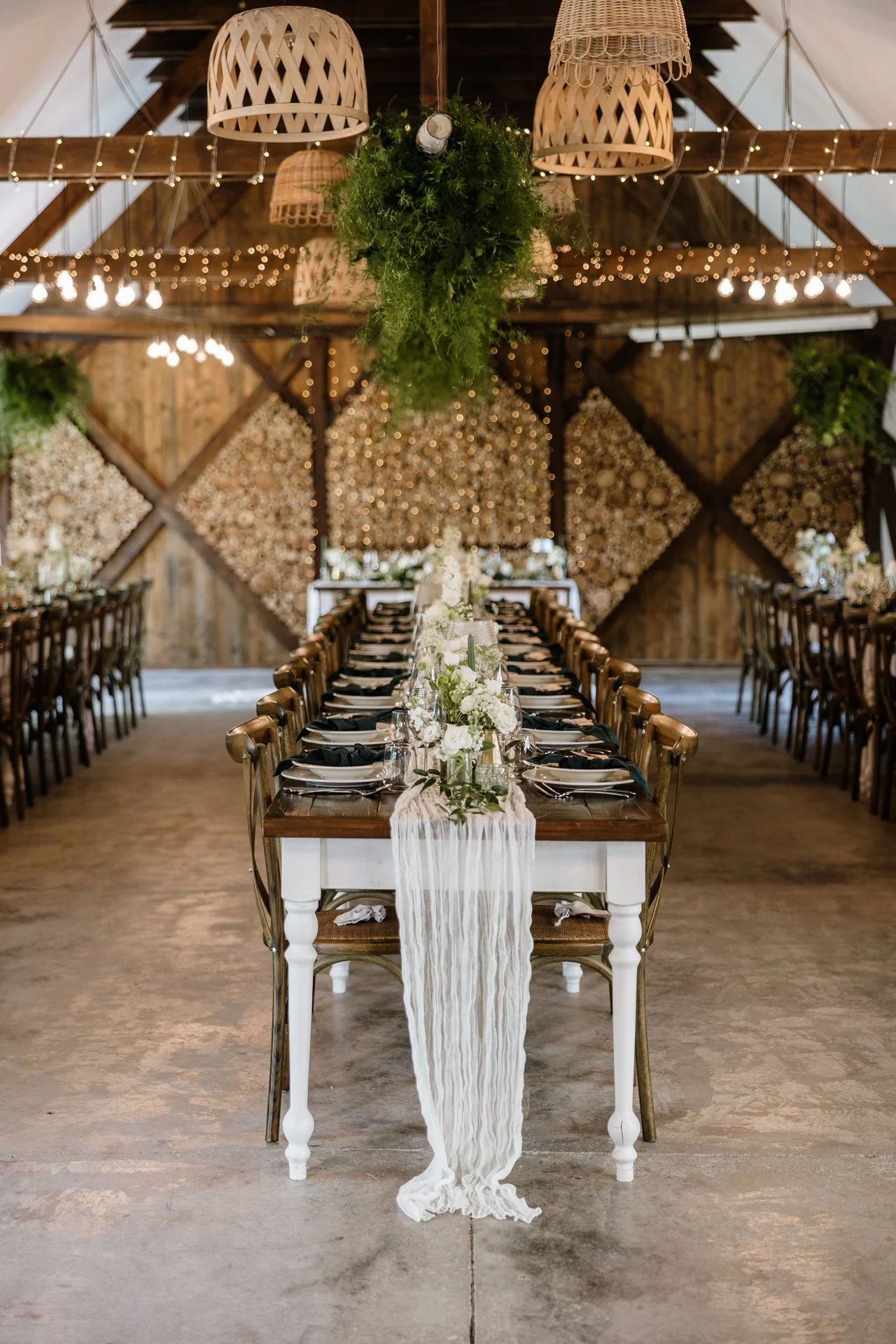 Elegant dining table set up in a rustic barn with flower arrangements, black napkins, and a white table runner. Decor includes greenery and string lights, with a backdrop of wooden walls and floral wall art.
