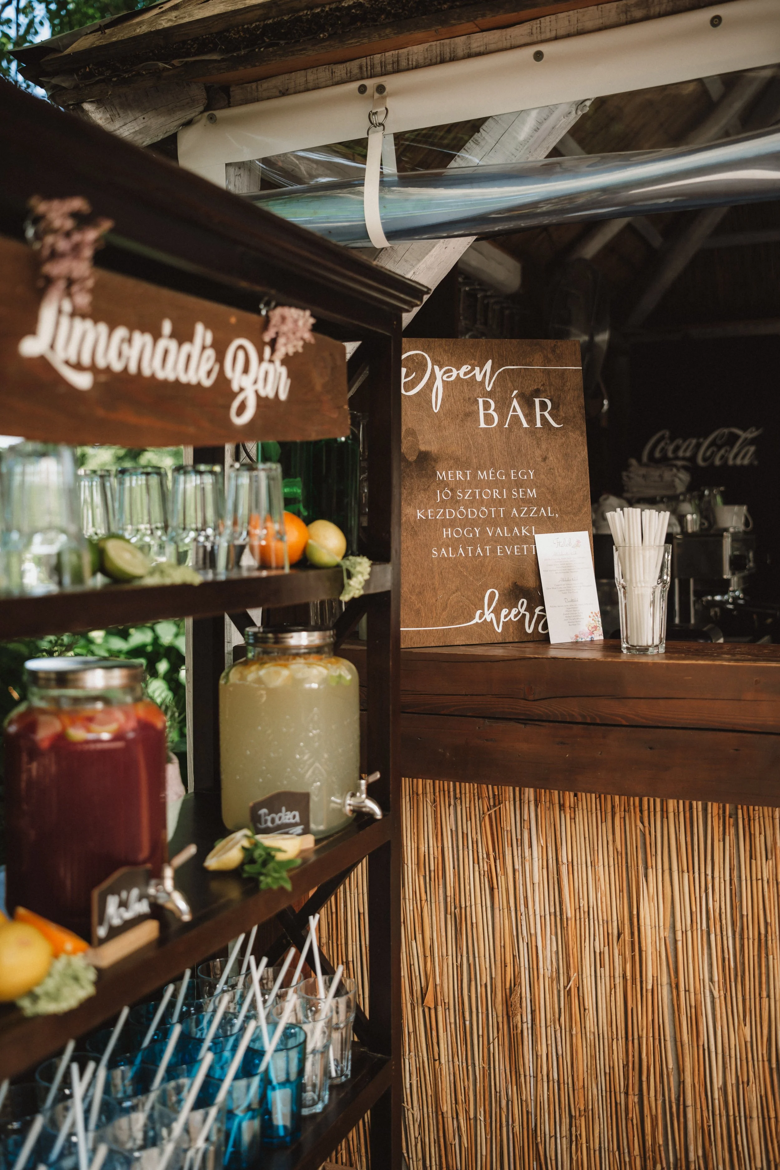 A beverage bar with jars of colorful drinks, lemons, limes, and garnishes, and signs in Hungarian and English indicating that it is an open bar.