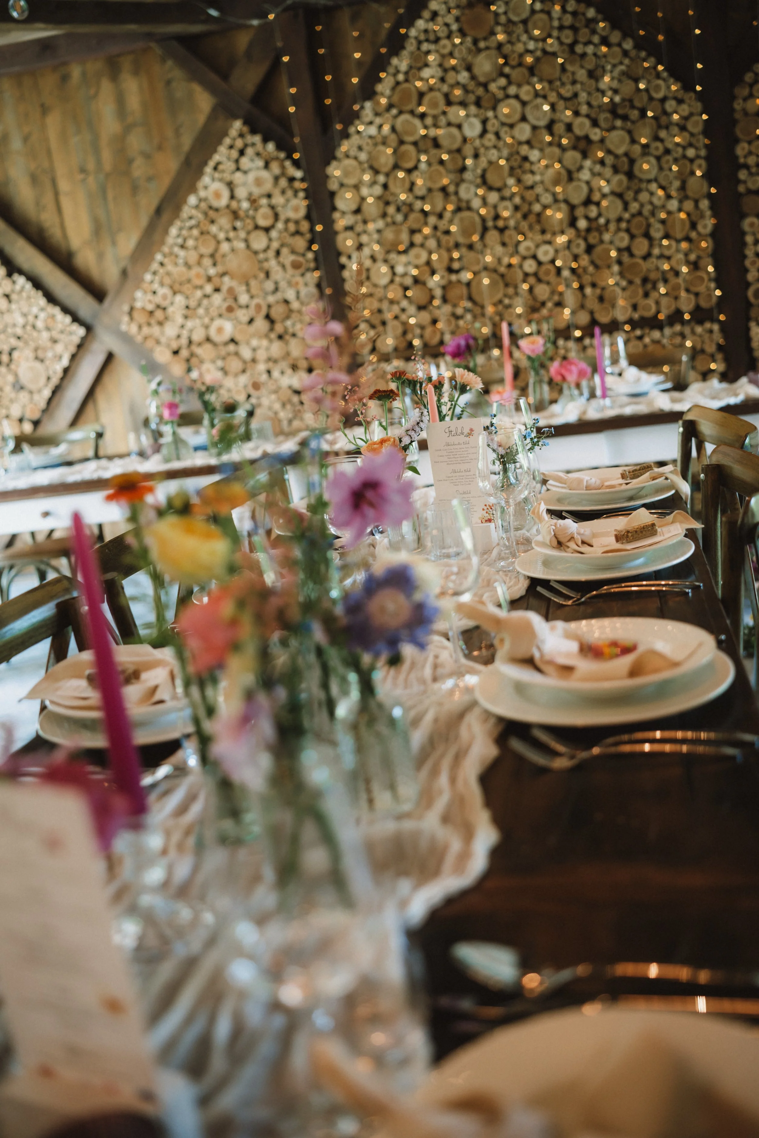A decorated banquet table in a rustic setting with floral centerpieces, candles, and place settings, with a wood log wall backdrop and string lights.