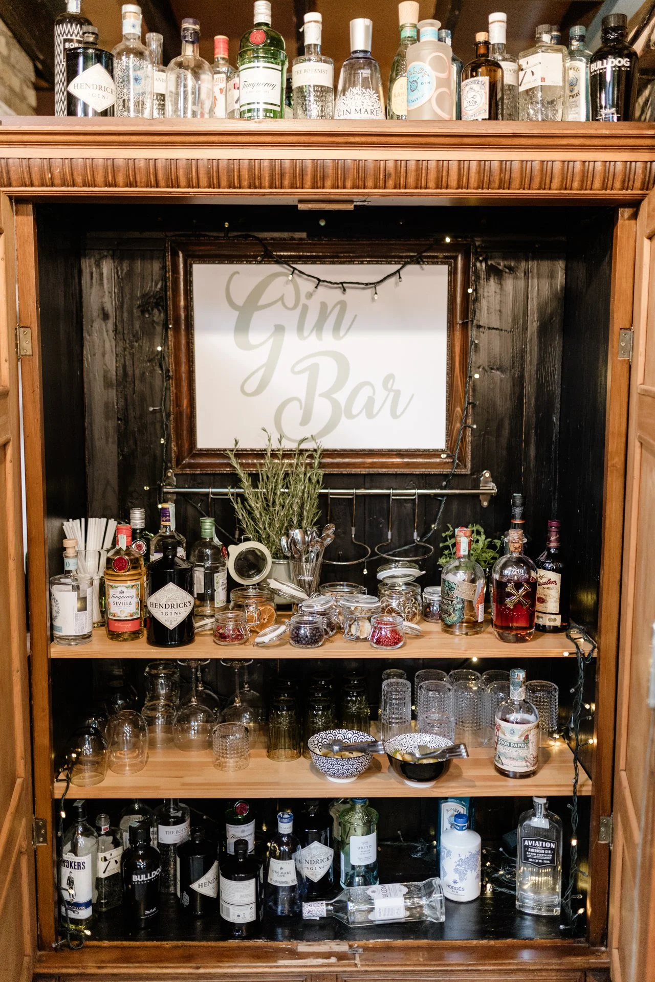 A wooden bar cabinet with a collection of gin bottles, glasses, jars of berries, a bowl, and a small potted herb plant. A sign reading 'Gin Bar' is in the center, decorated with string lights.