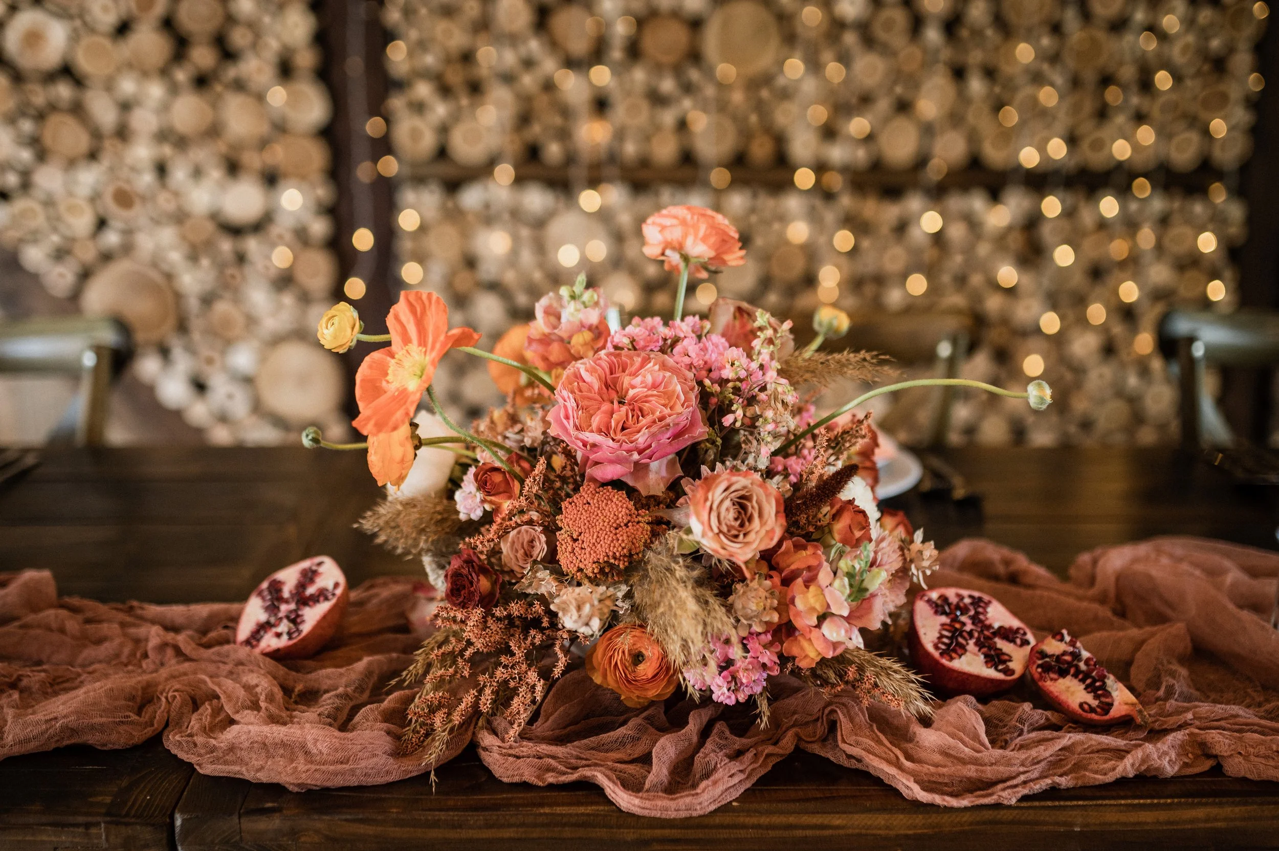 Virágdísz rózsaszín és barack színű virágokkal sötét fa asztalon, háttérben barna anyag és díszkövek.
