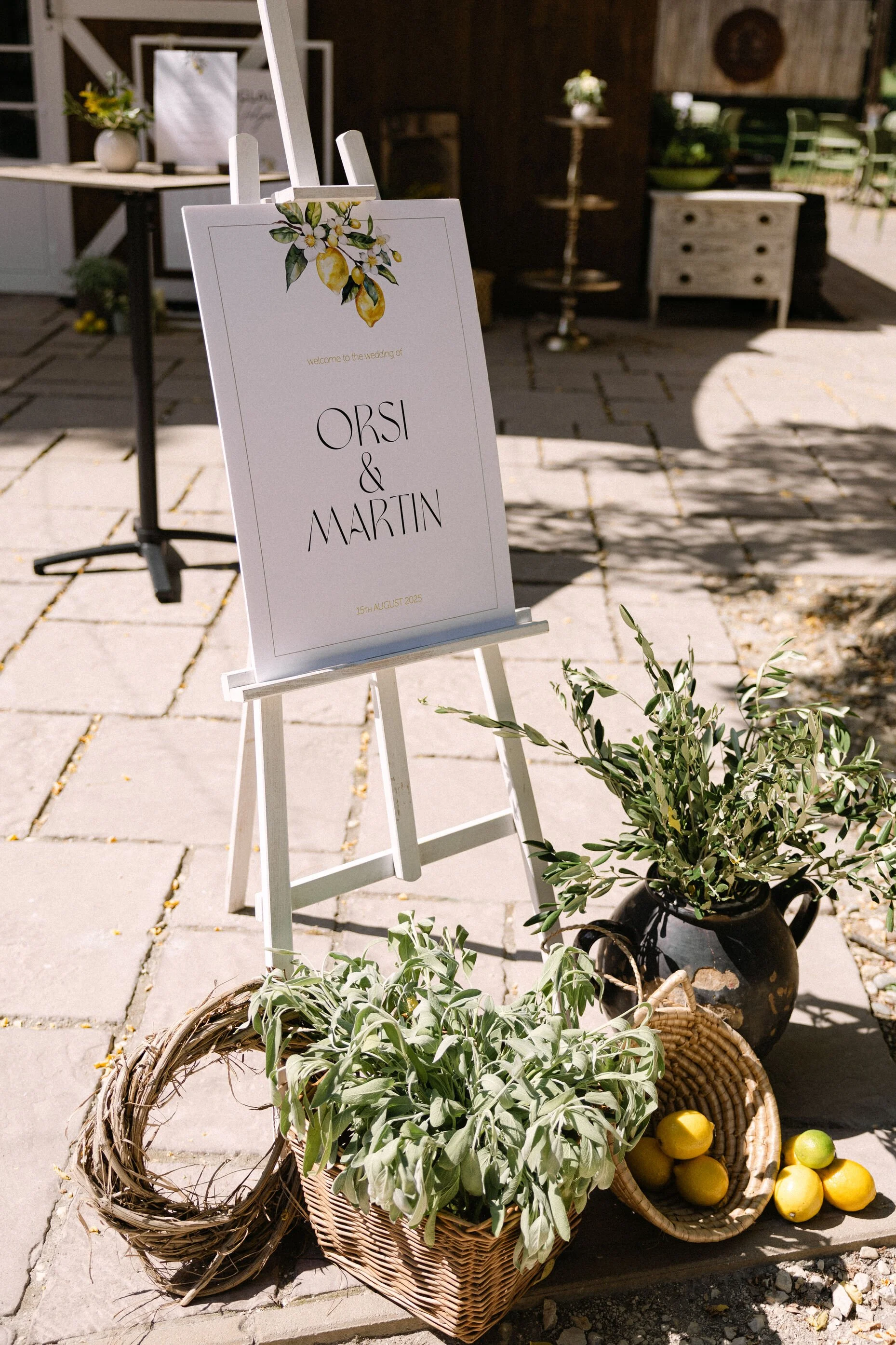 Esküvői tábla állványon az „Orsi & Martin” nevekkel és a „2025. augusztus 15.” dátummal, teraszon elhelyezve, citromokkal és zöld növényzettel díszítve, mellettük kosarak citrommal és zöld levelekkel.
