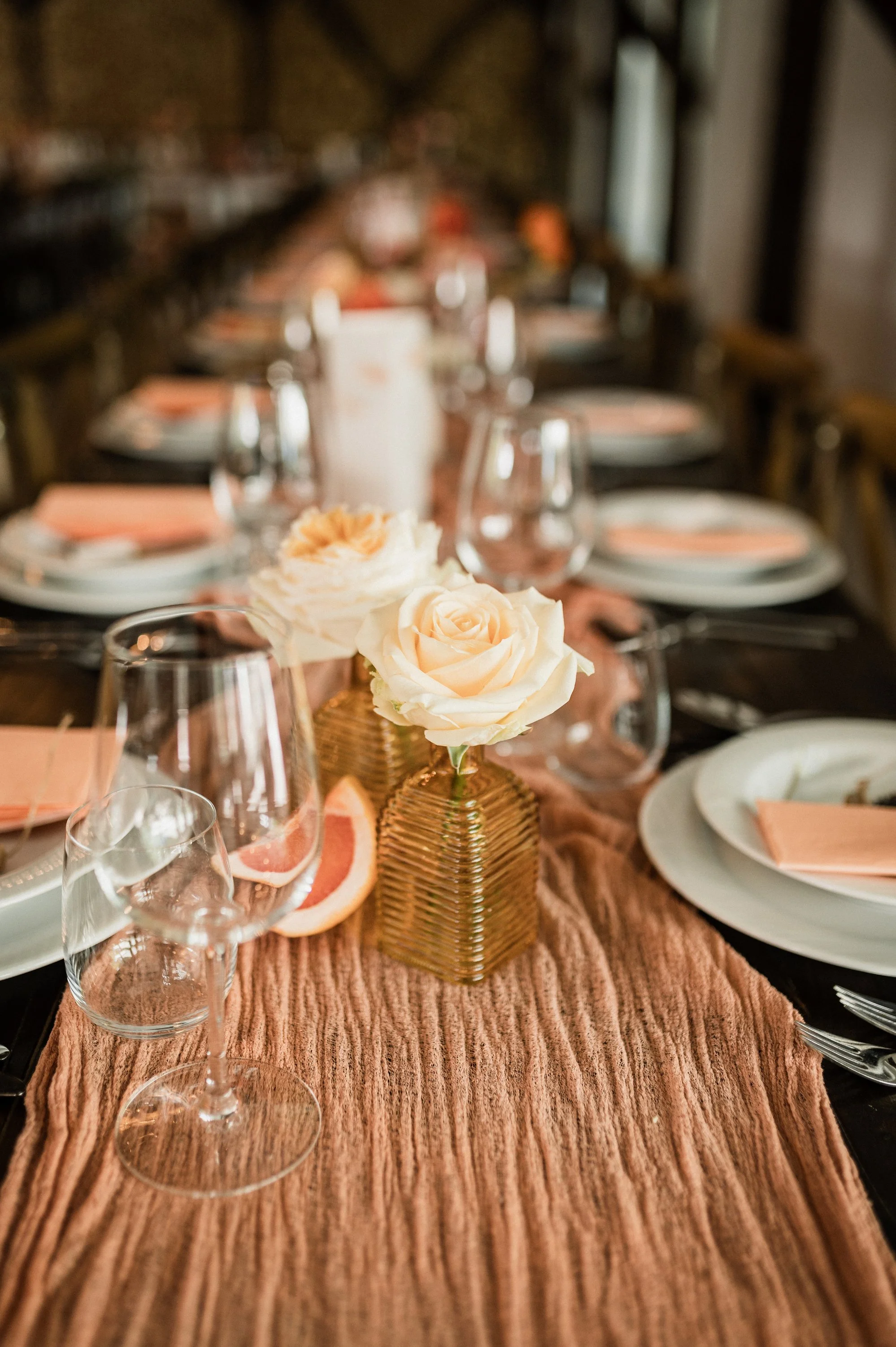 A dining table set for a special occasion with a ruffled pink table runner, white roses in gold vases, grapefruit slices, wine glasses, and white plates with pink napkins, in a cozy, rustic setting.