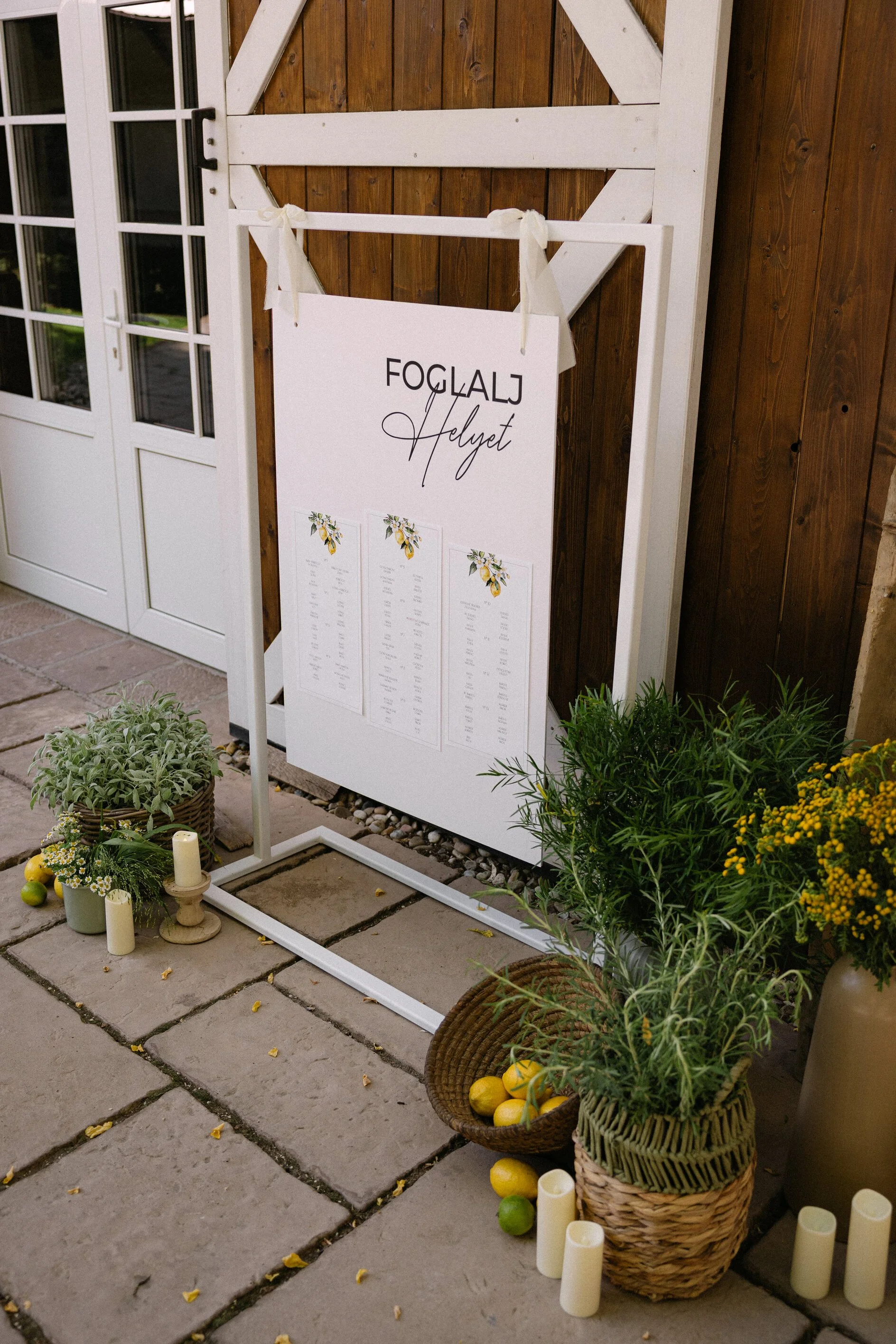 Wedding seating chart on a white sign with floral decorations, surrounded by yellow and green decorative elements and candles, outside a building with double glass doors and wooden walls.