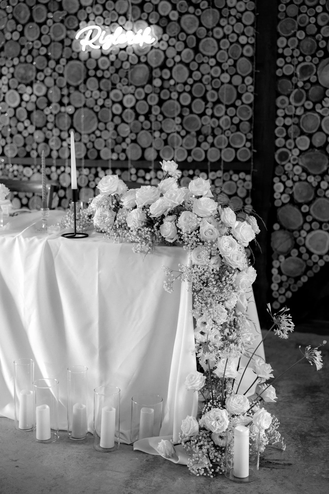 A decorated table with floral arrangements and candles in front of a wooden log wall, with a neon sign that reads 'Relax' in the background.