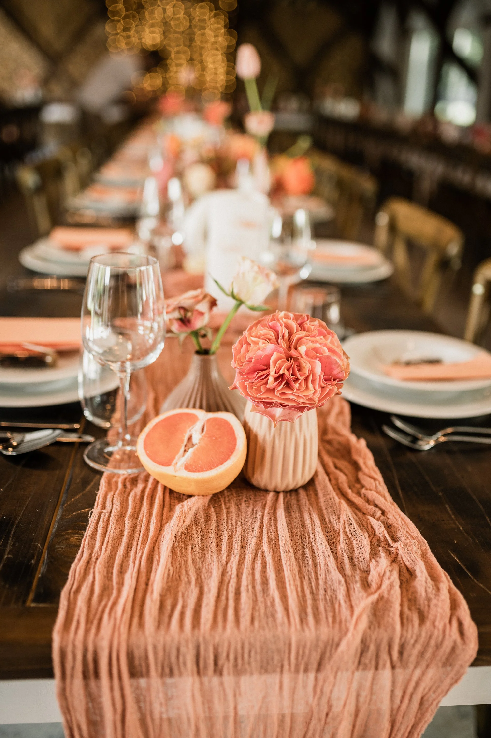 Díszített asztal rózsaszín virágokkal vázákban, szeletelt grapefruit-tal és pohár vízzel, megterítve tányérokkal, evőeszközökkel, szalvétákkal és rózsaszín asztali futóval, meleg, hangulatos környezetben.

