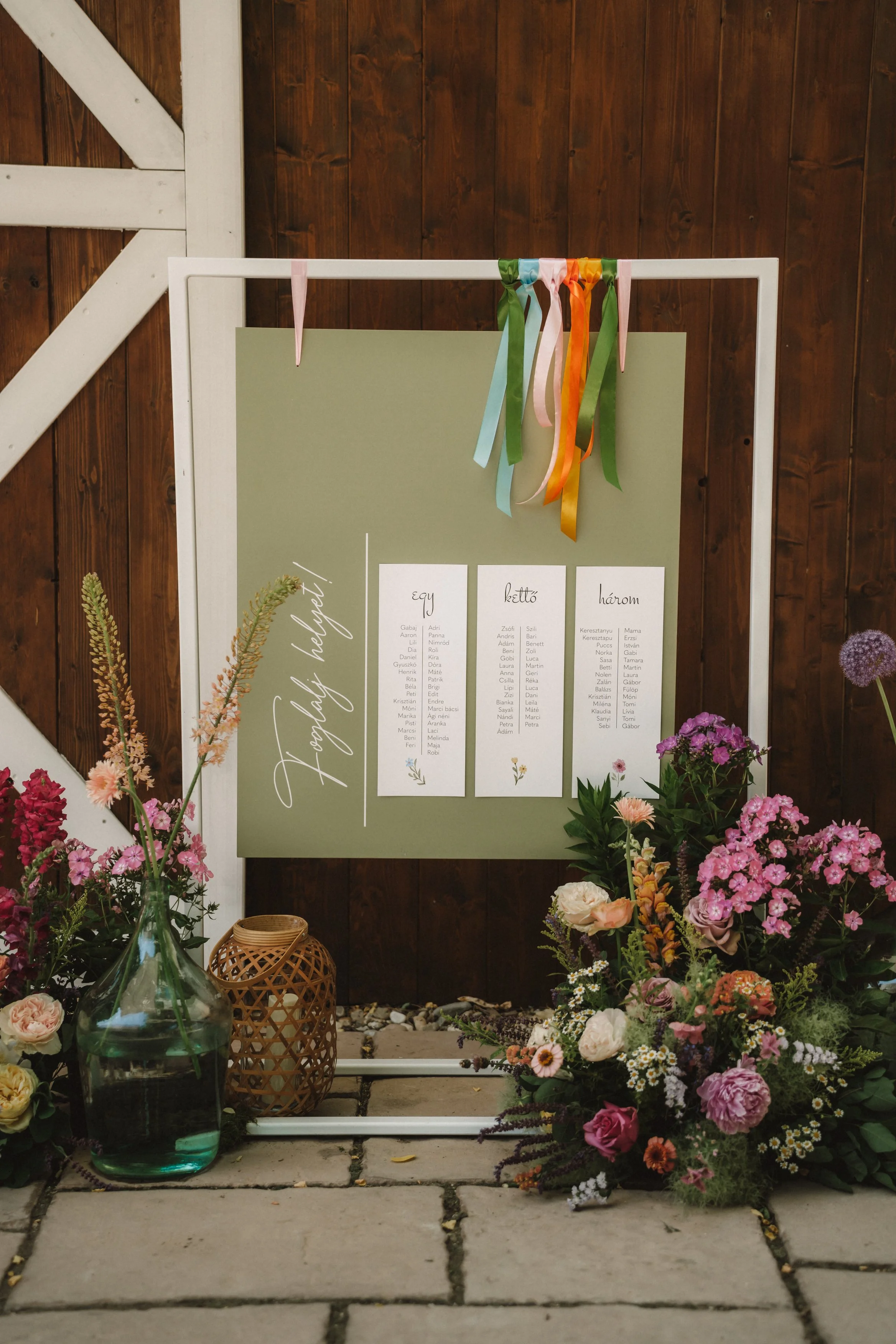Wedding seating chart displayed on a green board with white lettering, decorated with colorful ribbons, surrounded by flowers and vases on a stone surface against a dark wooden background.