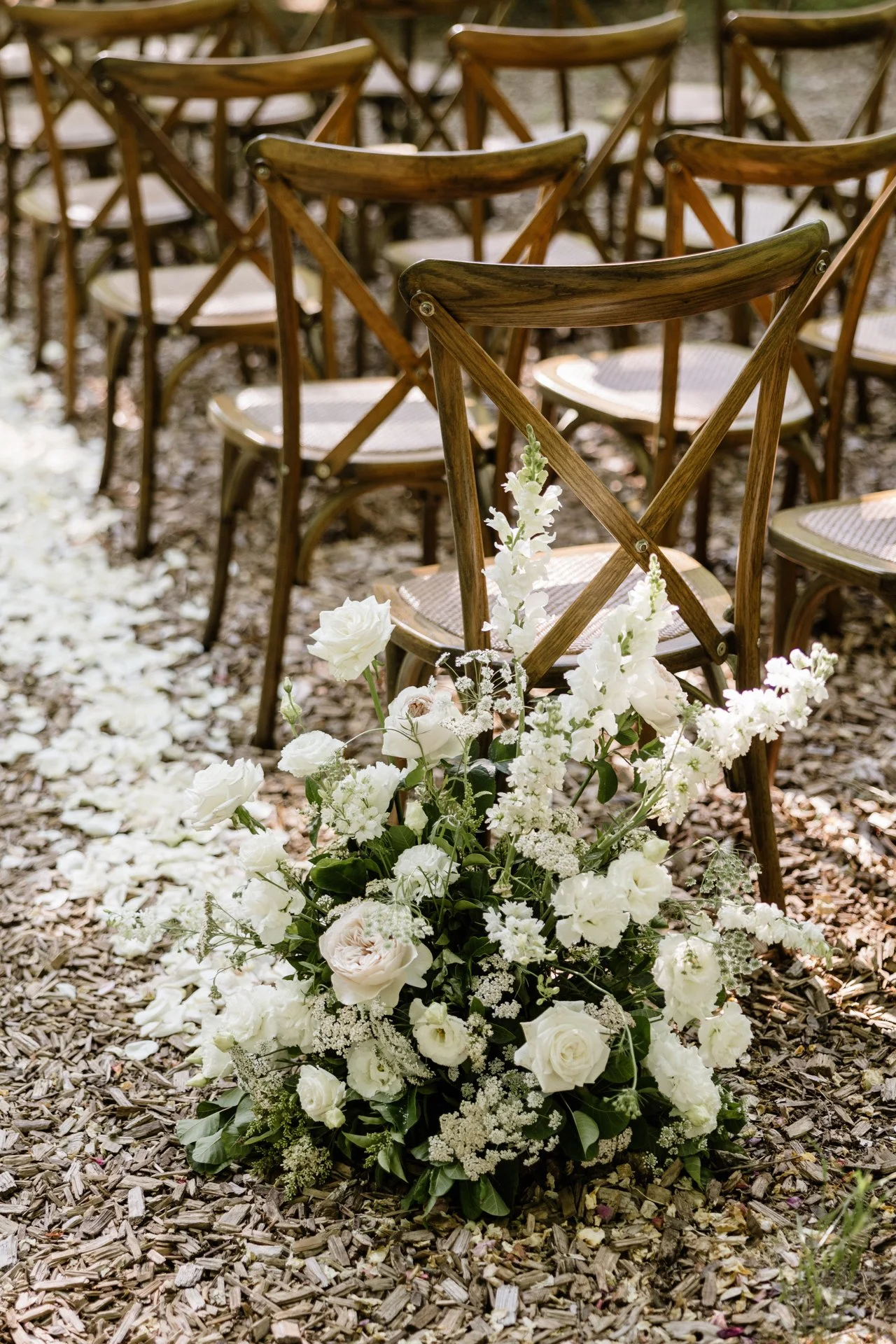 White floral arrangement with roses and other flowers placed in front of wooden chairs arranged for an outdoor event.