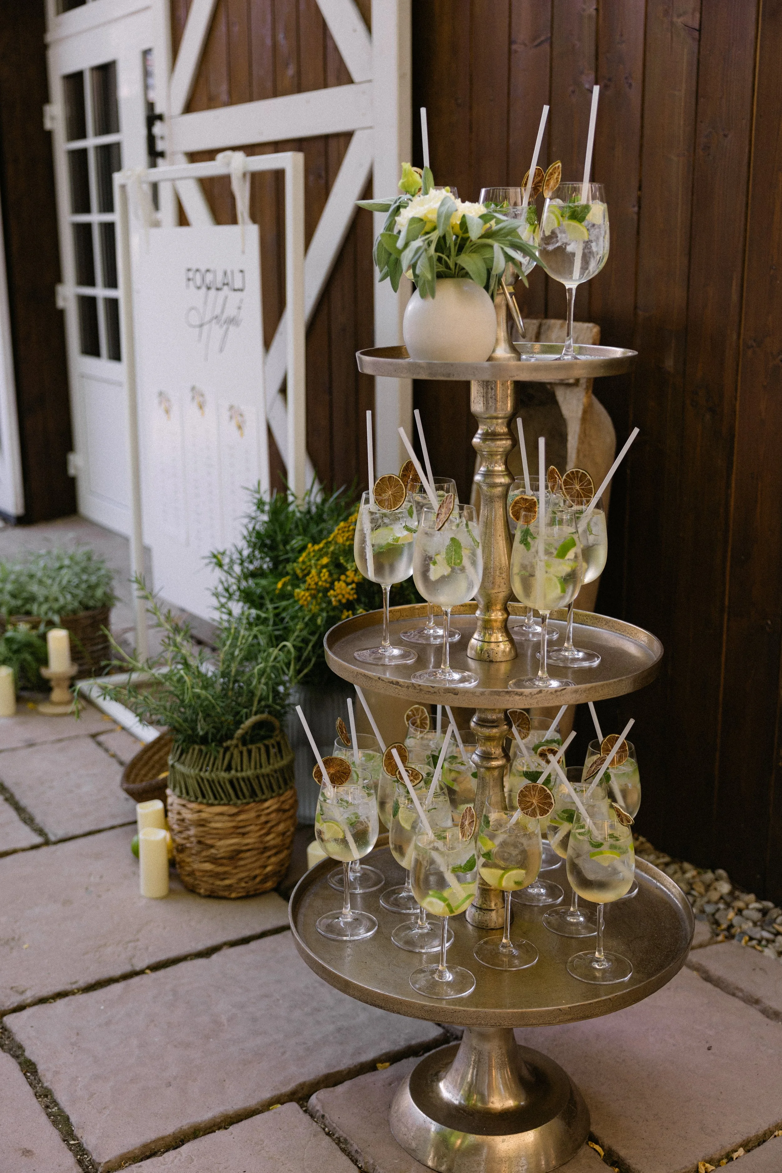 A three-tiered metallic stand with glasses of lemonade garnished with lemon slices and mint leaves, decorated with dried lemon slices and straws, set in an outdoor setting with a plant basket and candles nearby.