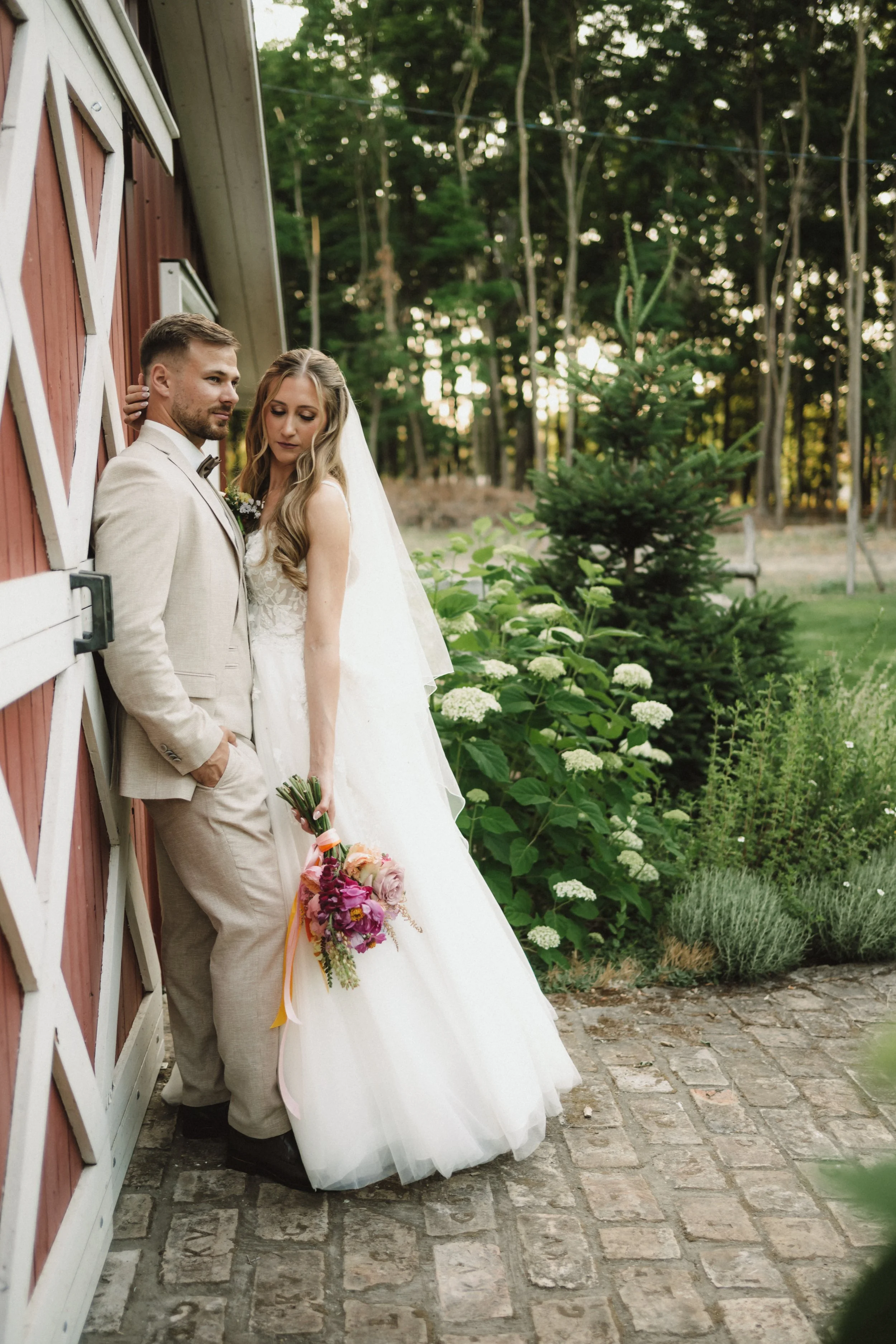 A bride and groom leaning against a red barn exterior, surrounded by greenery and white flowers, on their wedding day.