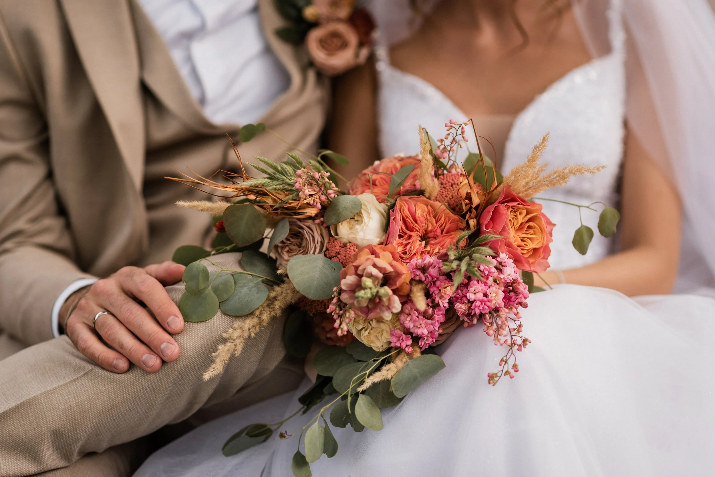 A menyasszony és a vőlegény egy esküvői ceremónia során egy csokrot tartanak kezükben, amely rózsaszín, narancs és krém színű virágokból, zöld növényzetből és szárított füvekből áll.
