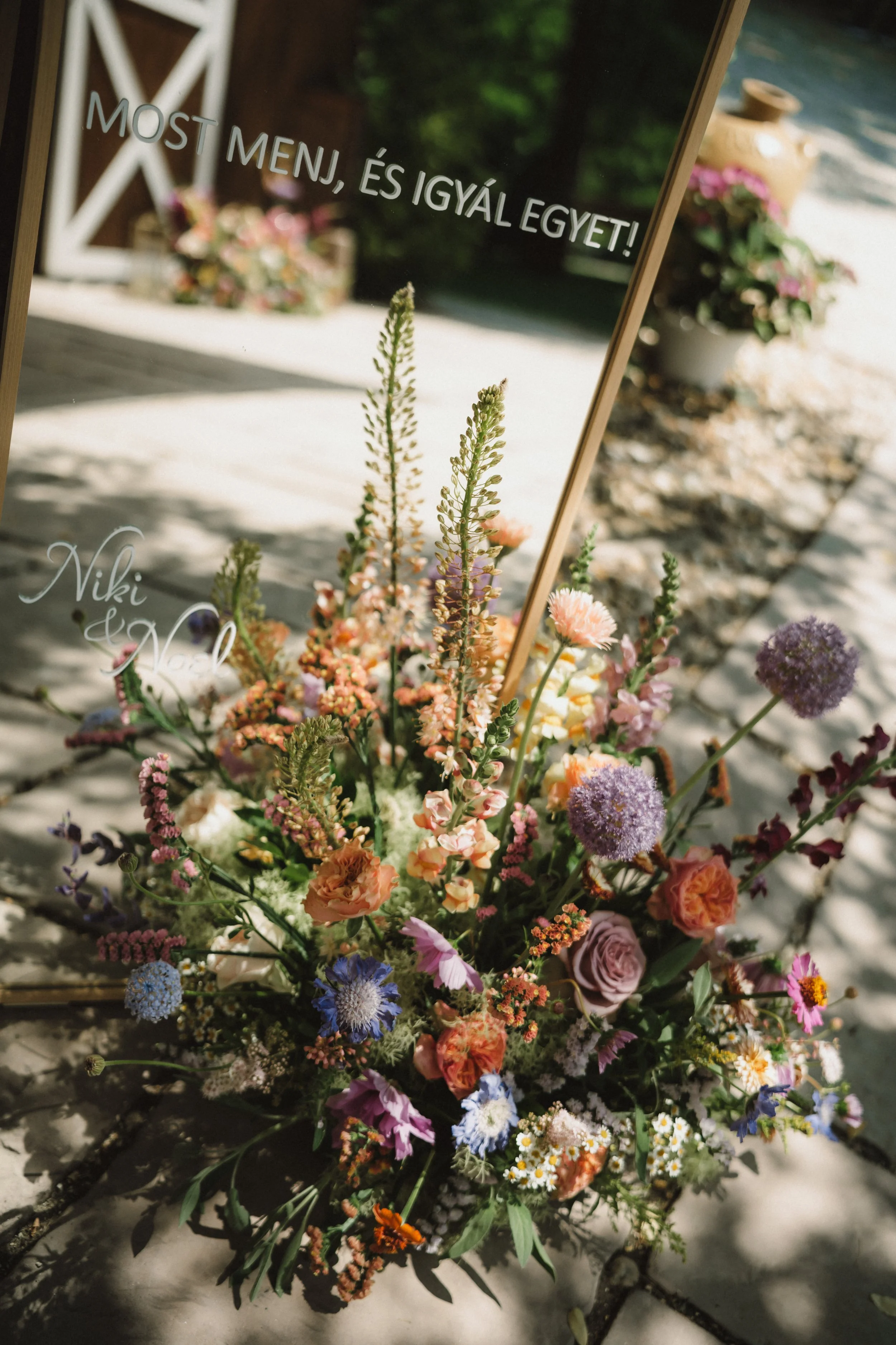 A floral arrangement with various colorful flowers placed on the ground outside, with a mirror that has Hungarian text and the name 'Niki & Daniel' written on it.