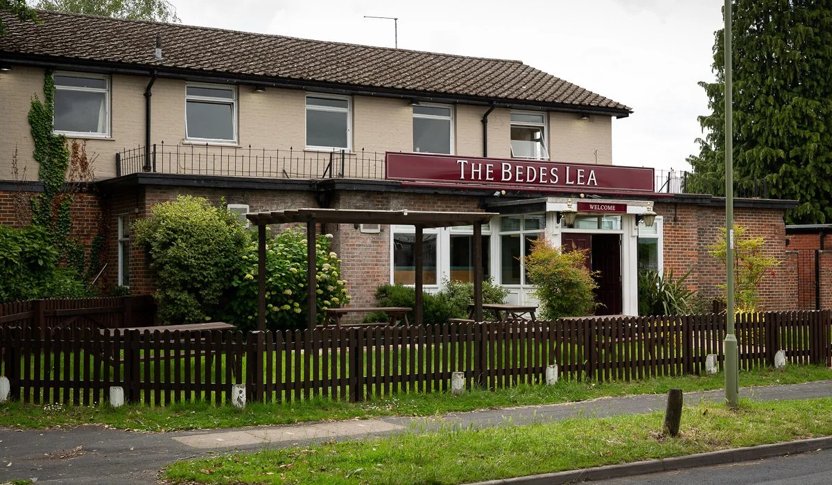 A two-story building with a sign that reads 'The Bedes Lea,' featuring a red and white color scheme, a small front garden with bushes and a wooden fence, and a sidewalk and street in front.