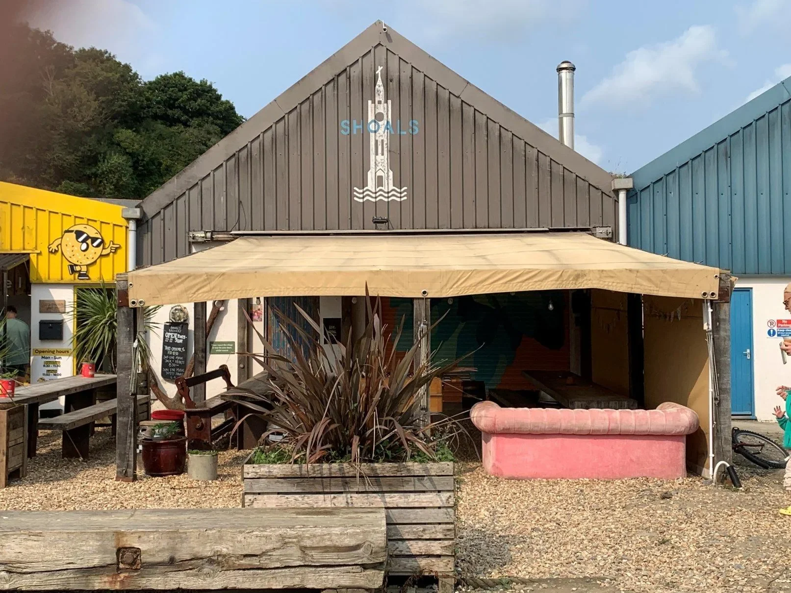 A rustic outdoor diner with a yellow and gray building, featuring a cartoon ice cream cone sign on the left and a blackboard menu near the entrance. The main building has a brown wooden exterior with a painted lighthouse and the word 'SHOALS' on top. A beige canopy covers part of the seating area, which includes a pink upholstered bench and plants in wooden planters. Gravel ground surrounds the area, and there are other colorful buildings nearby.