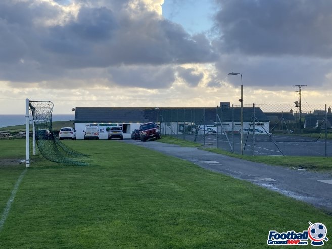 A grassy sports field with a soccer goal and a parking lot behind a chain-link fence. The sky is cloudy.