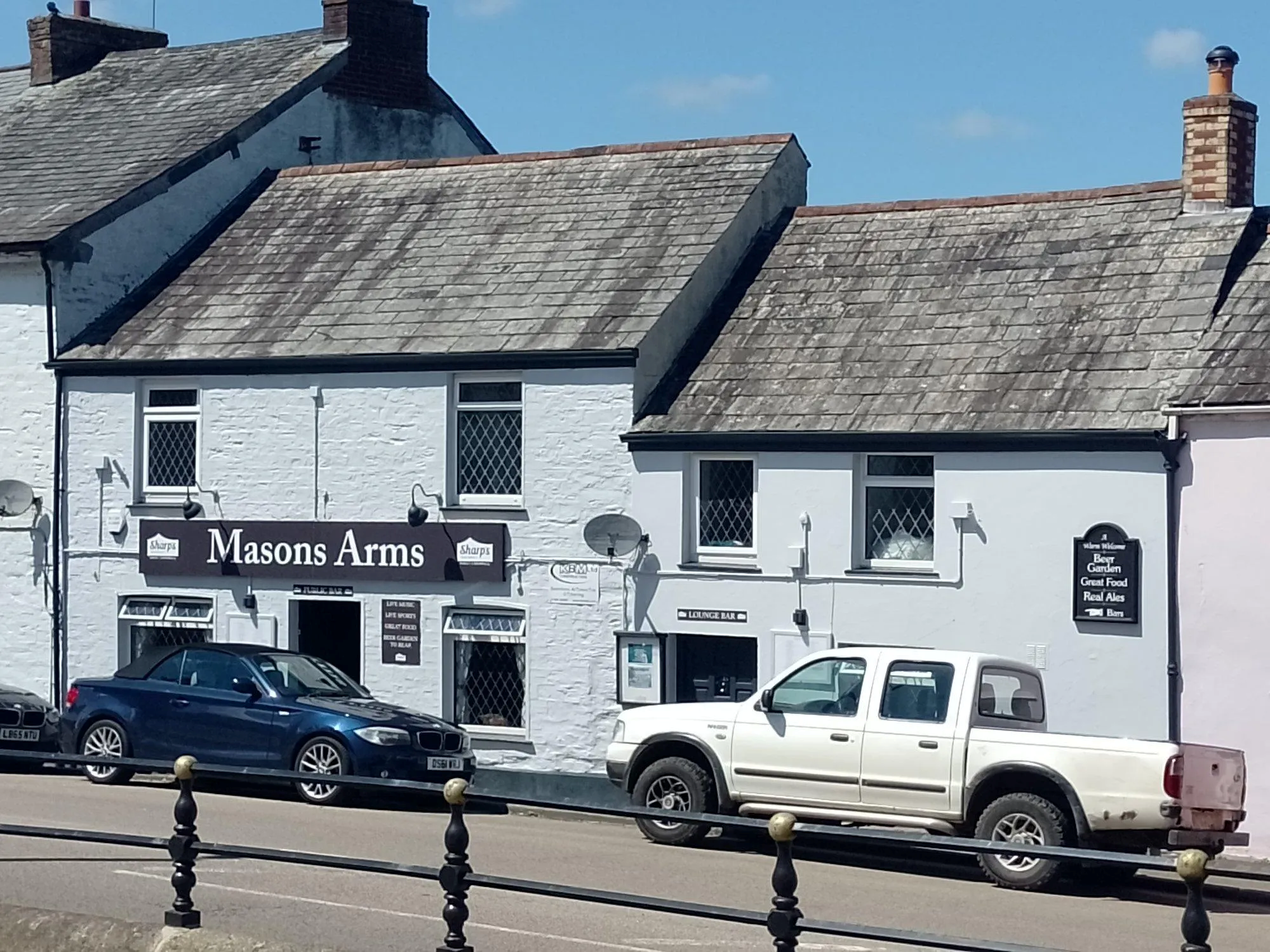 A row of white buildings with slate roofs, with a pub named Masons Arms on the left. The pub has a signboard. Two cars, a dark blue one and a white pickup truck, are parked in front.