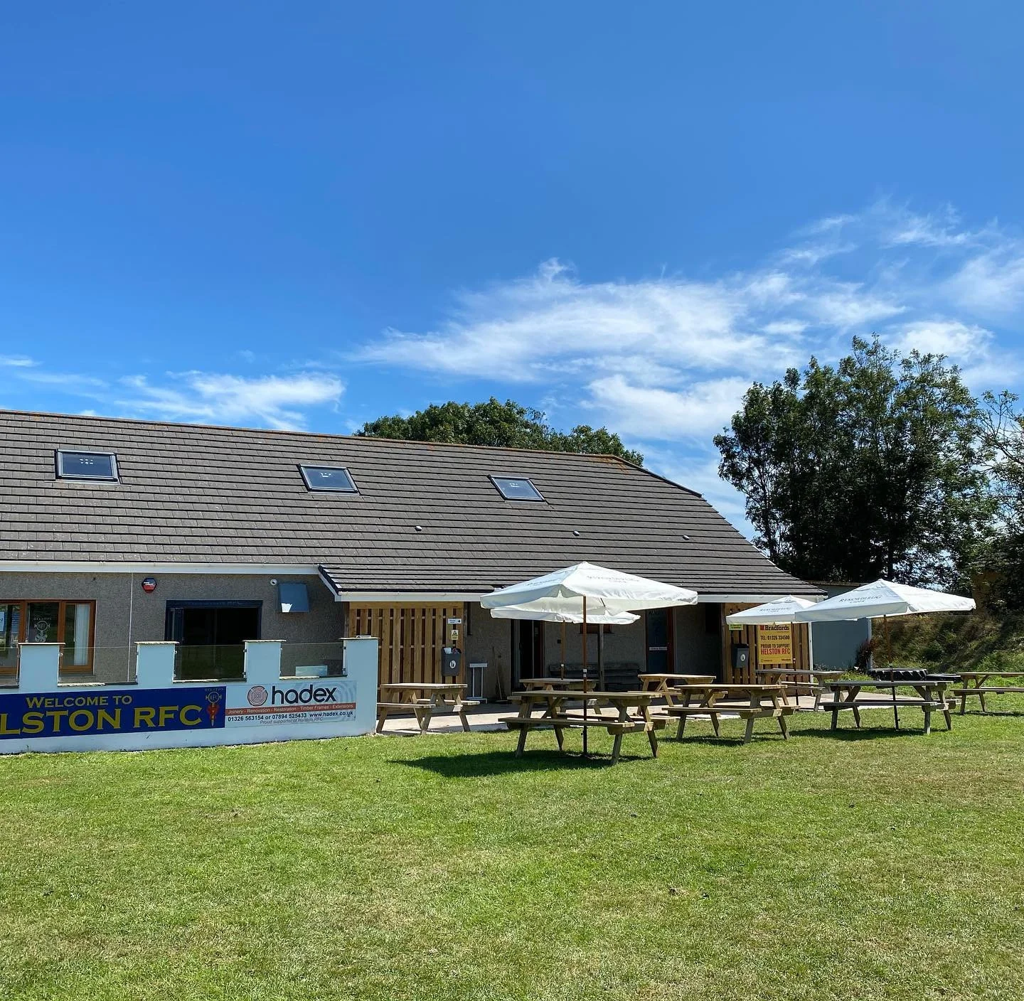 View of an outdoor seating area with picnic tables and umbrellas in front of a building with a sign that reads 'Welcome to Holston RFC' on a sunny day with blue sky and some clouds.