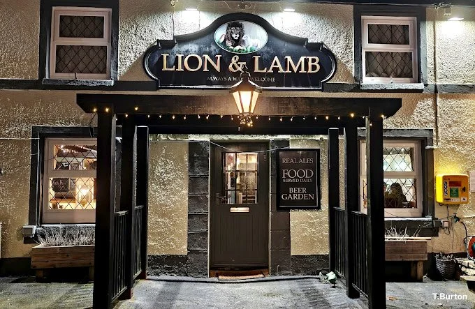 The exterior of a pub named 'Lion & Lamb' at dusk with outdoor seating, umbrellas, and string lights, featuring a central pathway, potted plants, and signs advertising food and drink.
