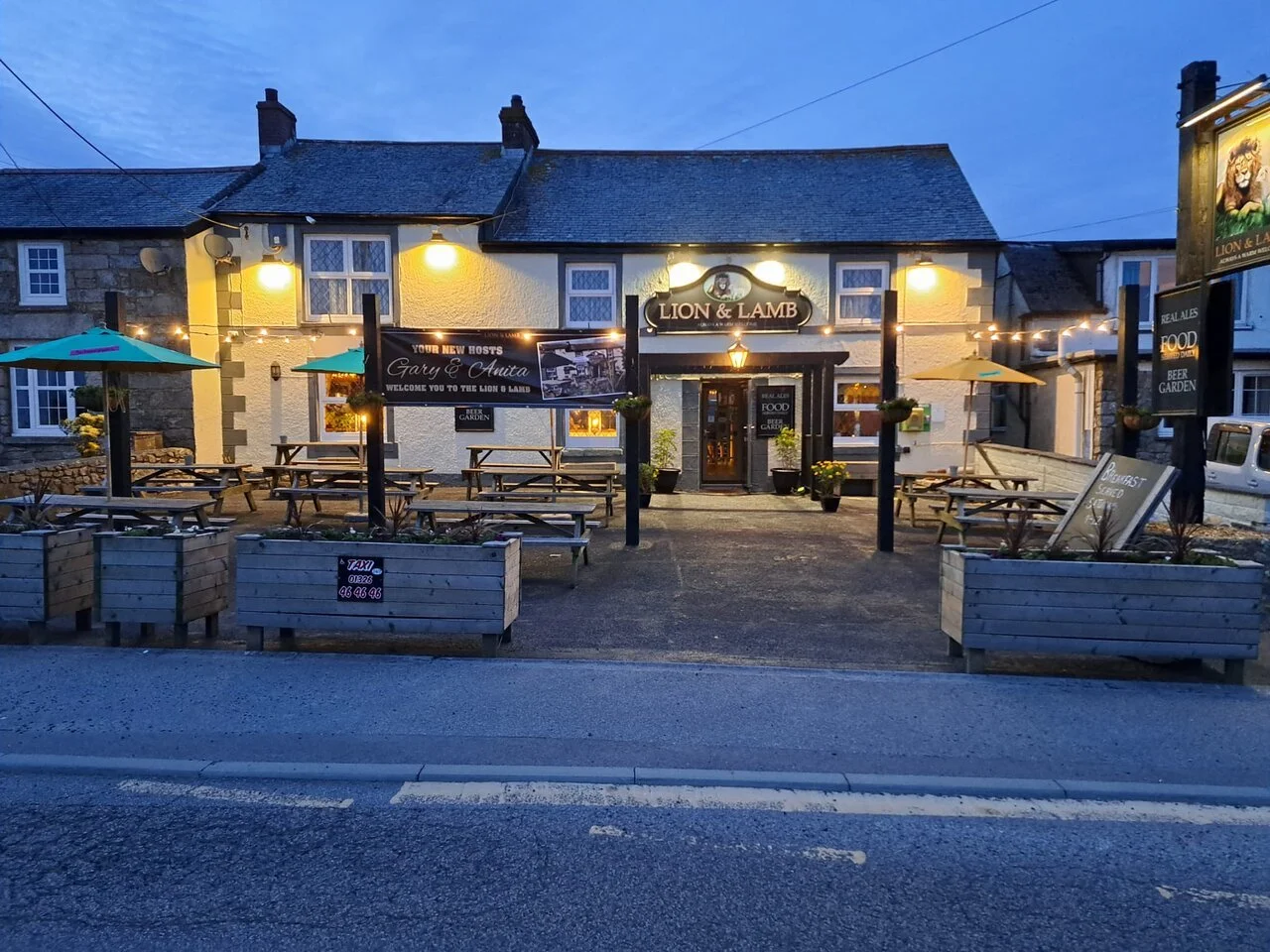 The exterior of a pub named 'Lion & Lamb' at dusk with outdoor seating, umbrellas, and string lights, featuring a central pathway, potted plants, and signs advertising food and drink.
