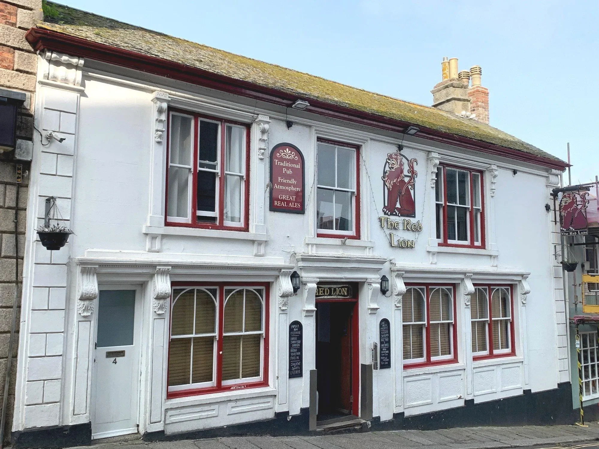 Exterior of The Red Lion pub, a white two-story building with red window frames and decorative white trim, featuring a lion illustration and signs advertising traditional pub atmosphere and great ales.