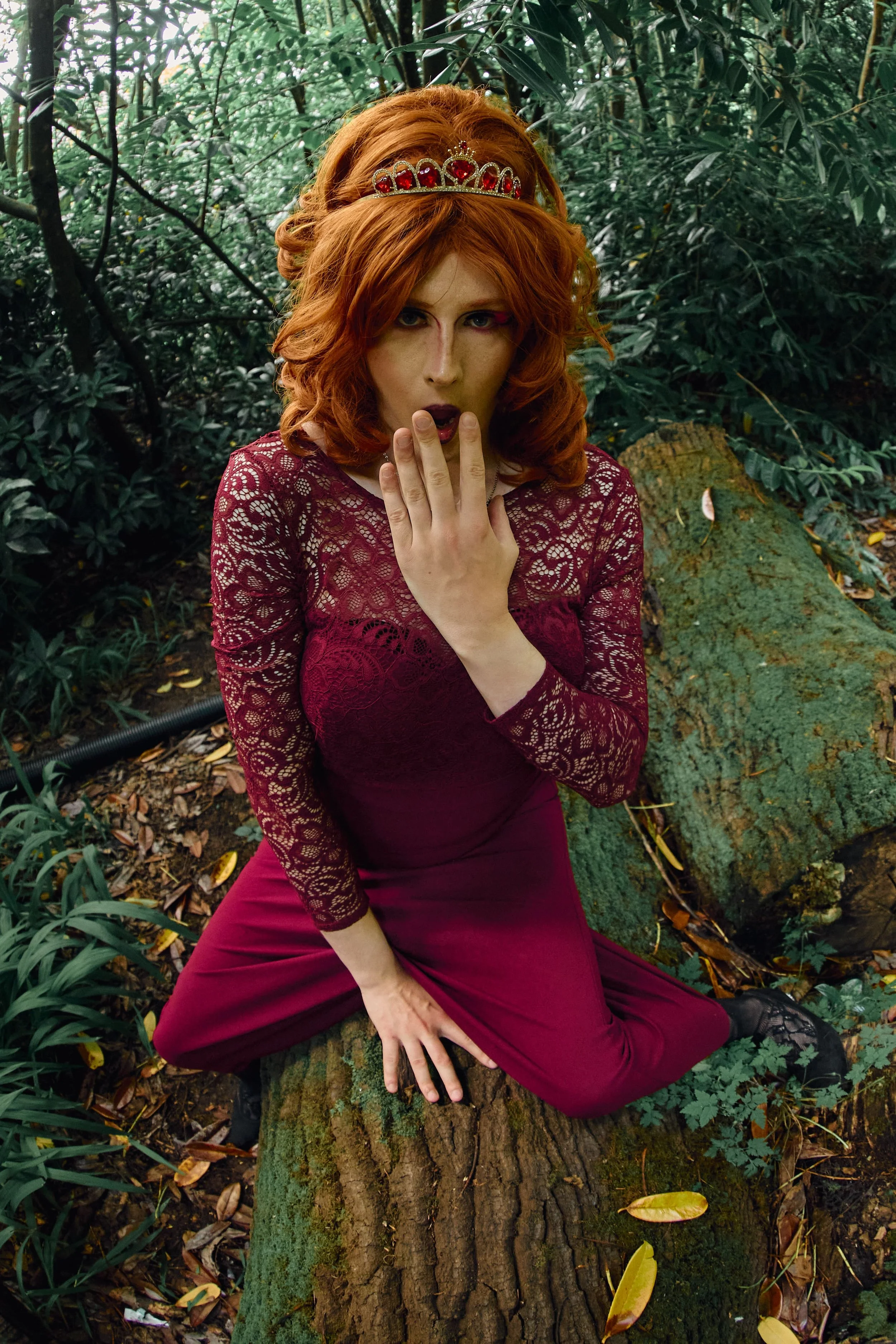 A woman with red hair, wearing a metal tiara, a burgundy lace top, and burgundy pants, sitting on a tree trunk in a forested area with green foliage around her.