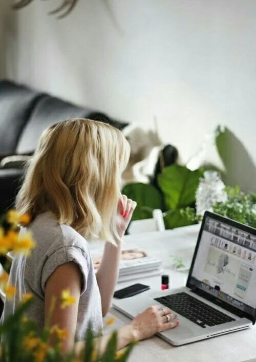 Een meisje met blond haar kijkt naar een laptop op een tafel, omringd door planten en bloemen in een woonkamer.