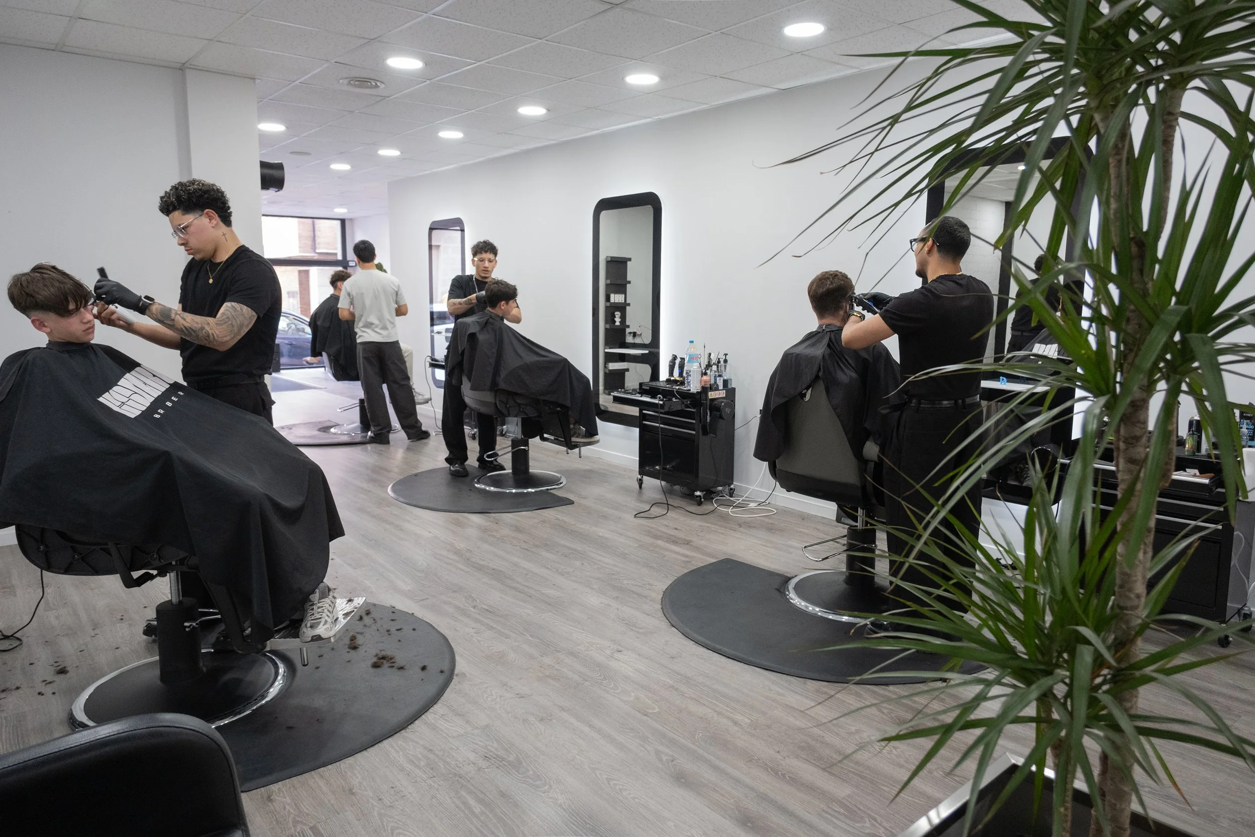 Salón de peluquería con varias personas trabajando y cortando cabello, sillas de estilista, espejos y productos de peinado en medio de un ambiente moderno con paredes blancas y mucha luz natural.
