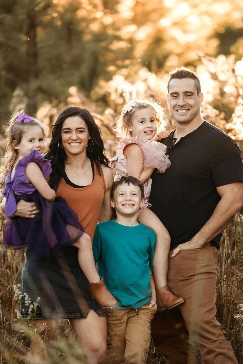 A family of five smiles outdoors during golden hour, with trees and sunlight in the background. The mother holds a young girl dressed in purple, another girl sits on the father's shoulder wearing pink, and a young boy stands in front wearing a teal shirt. The father is standing with one hand in his pocket, and the family appears happy and relaxed.