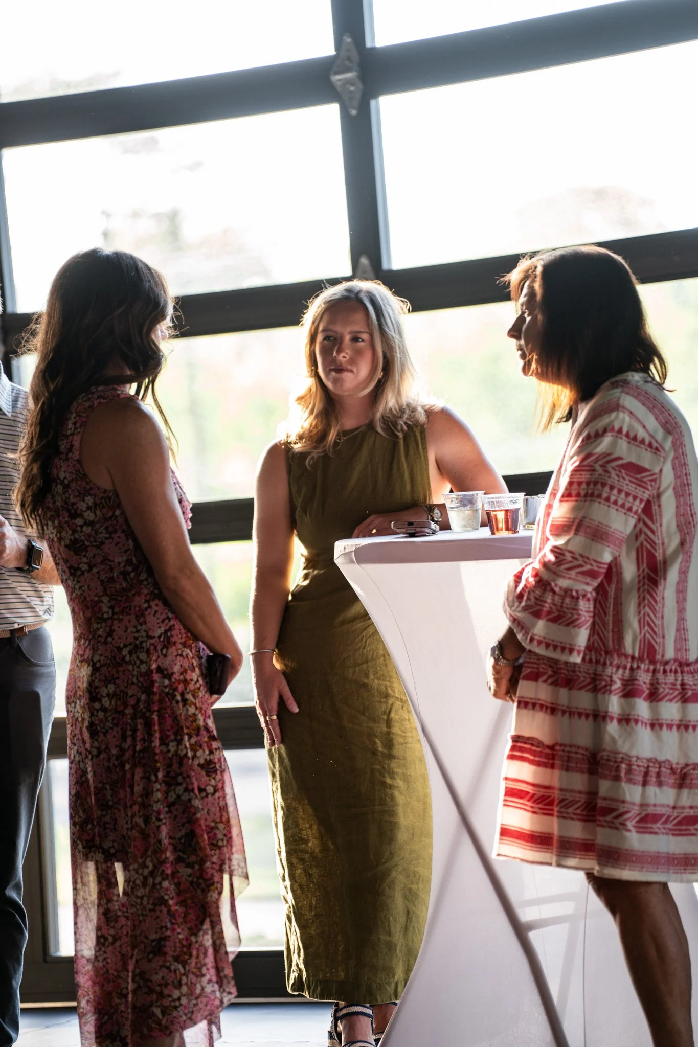 Four women talking at a round high-top table. They are indoors near large windows, with sunlight coming through.