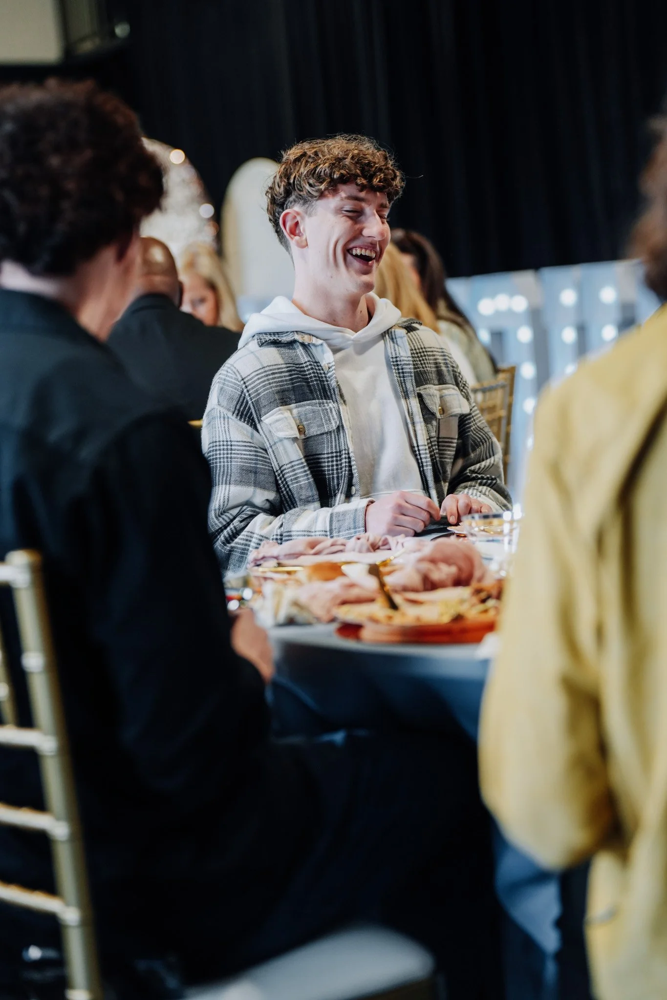 A young man with curly hair laughing and sitting at a table with food, surrounded by other people.