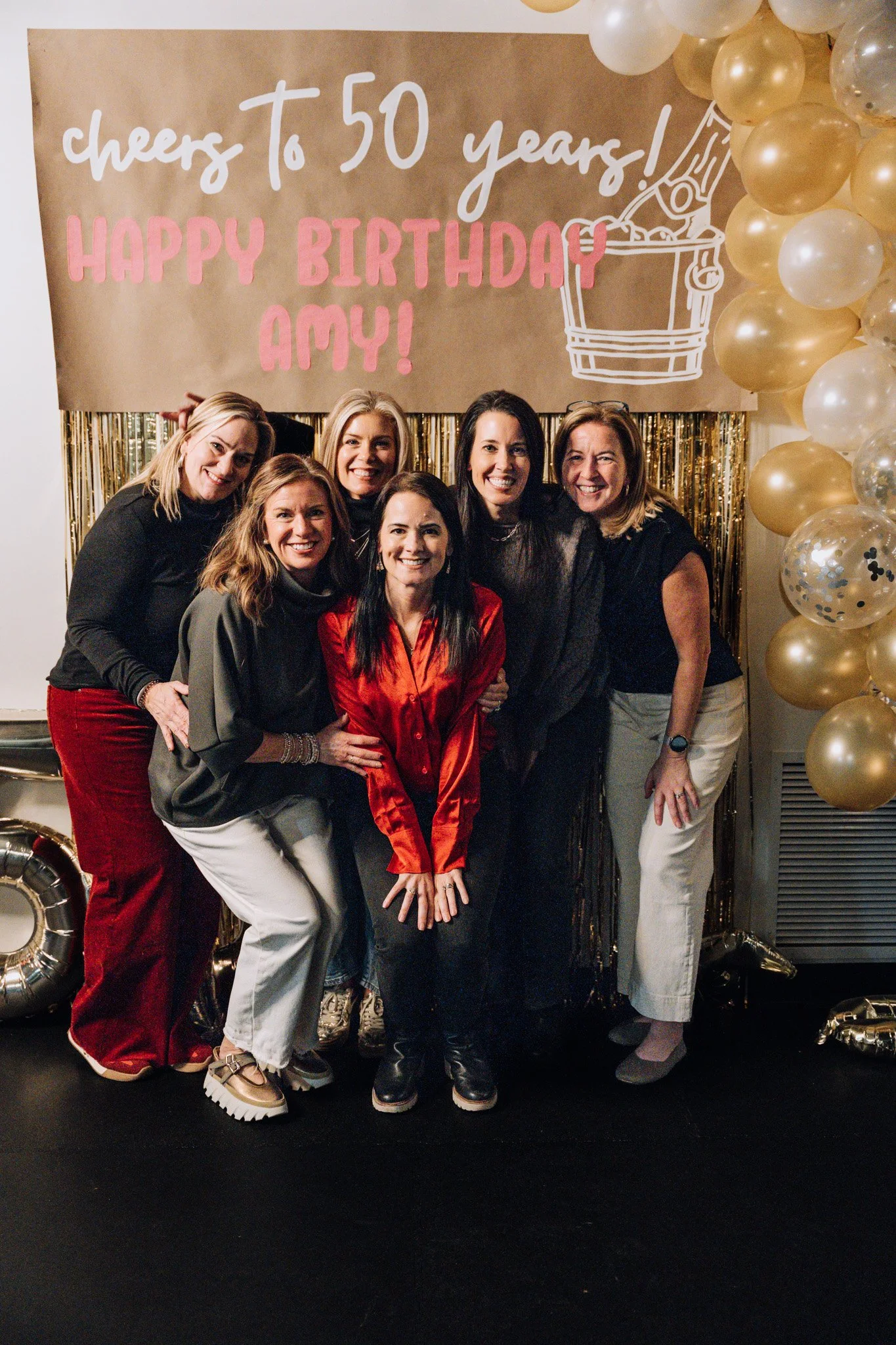 Group of seven women smiling at a celebration for Amy's 50th birthday, decorated with a gold backdrop, balloons, and a sign that says 'Cheers to 50 years! Happy birthday Amy!'.