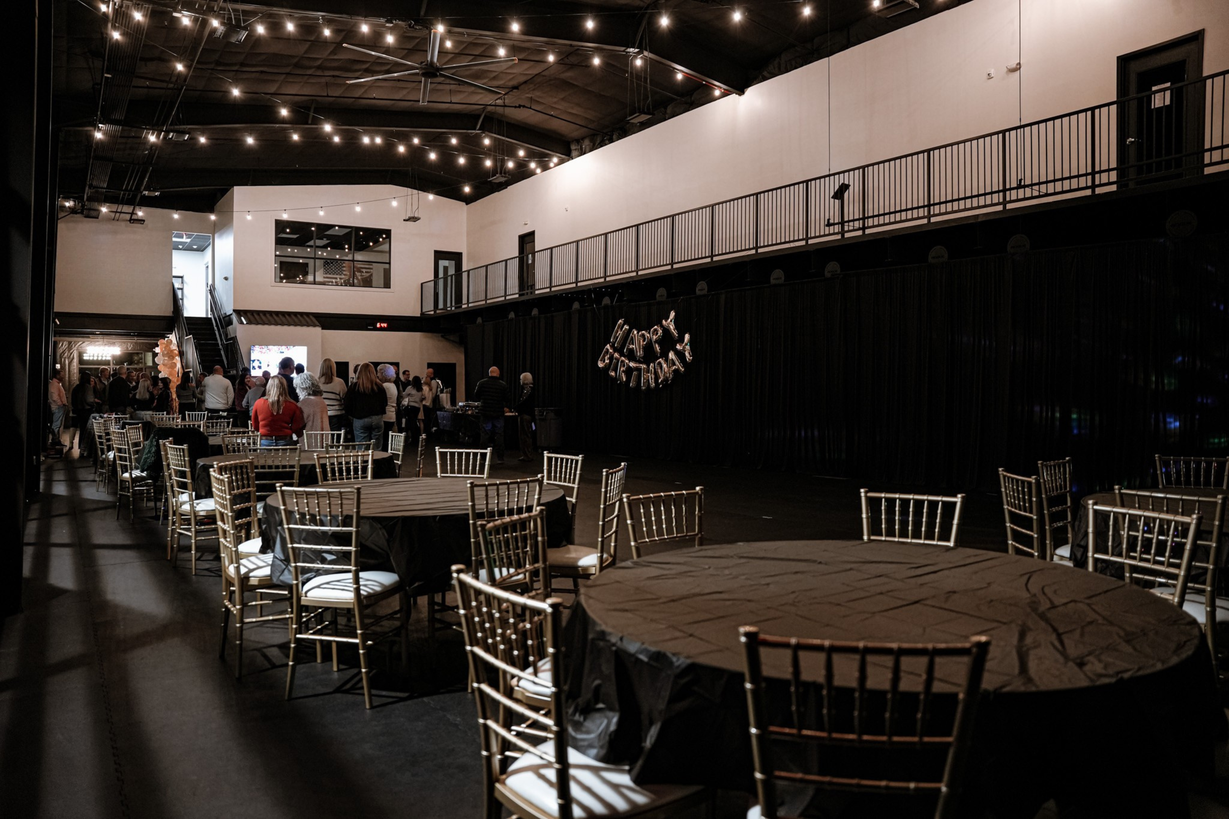 Wedding reception hall decorated with balloons and a 'Happy Birthday' banner, with people gathering near the tables and a DJ booth in the background.