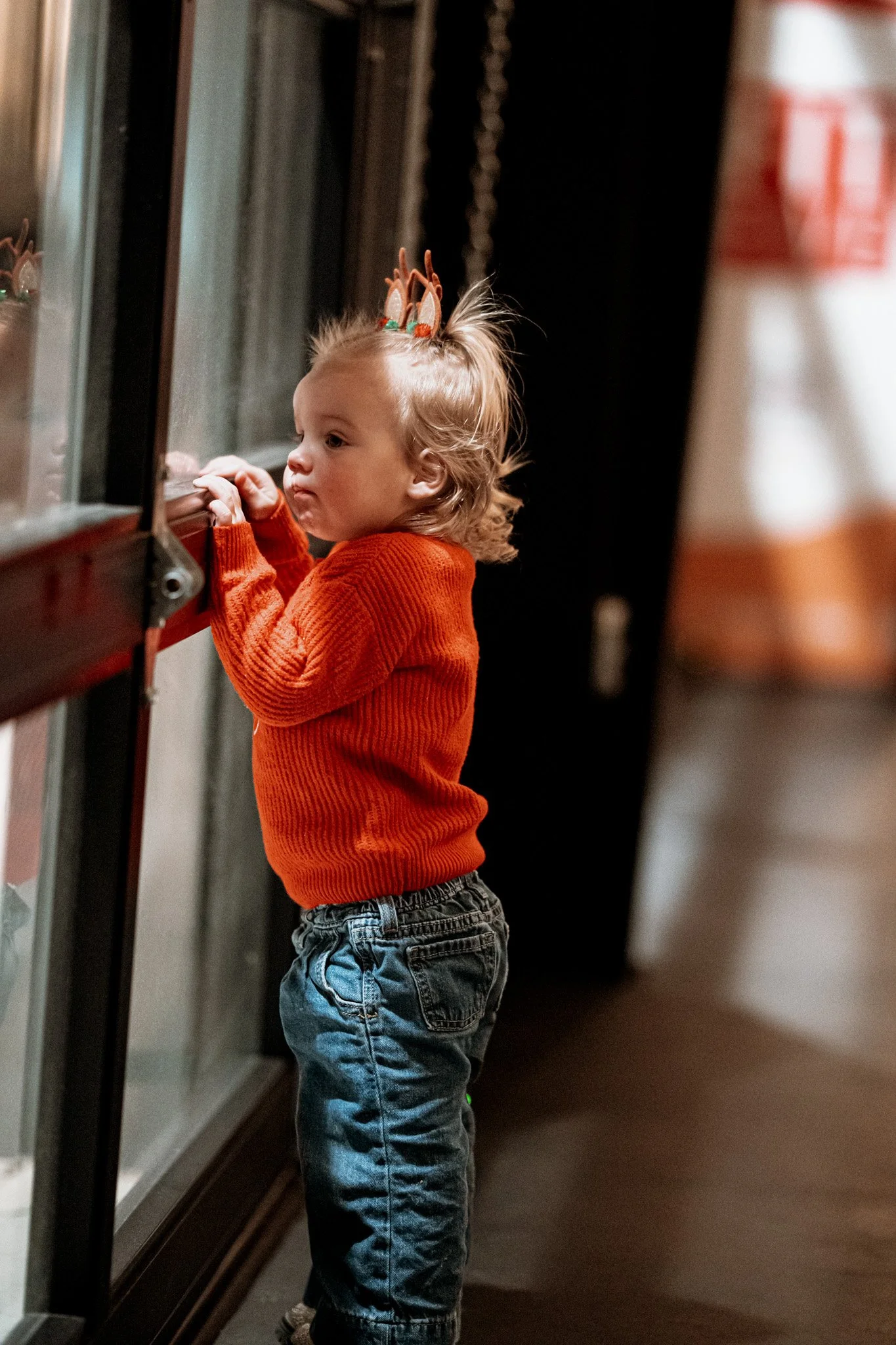 A young child with blonde hair wearing a reindeer headband, an orange sweater, and blue jeans looking through a glass display case.