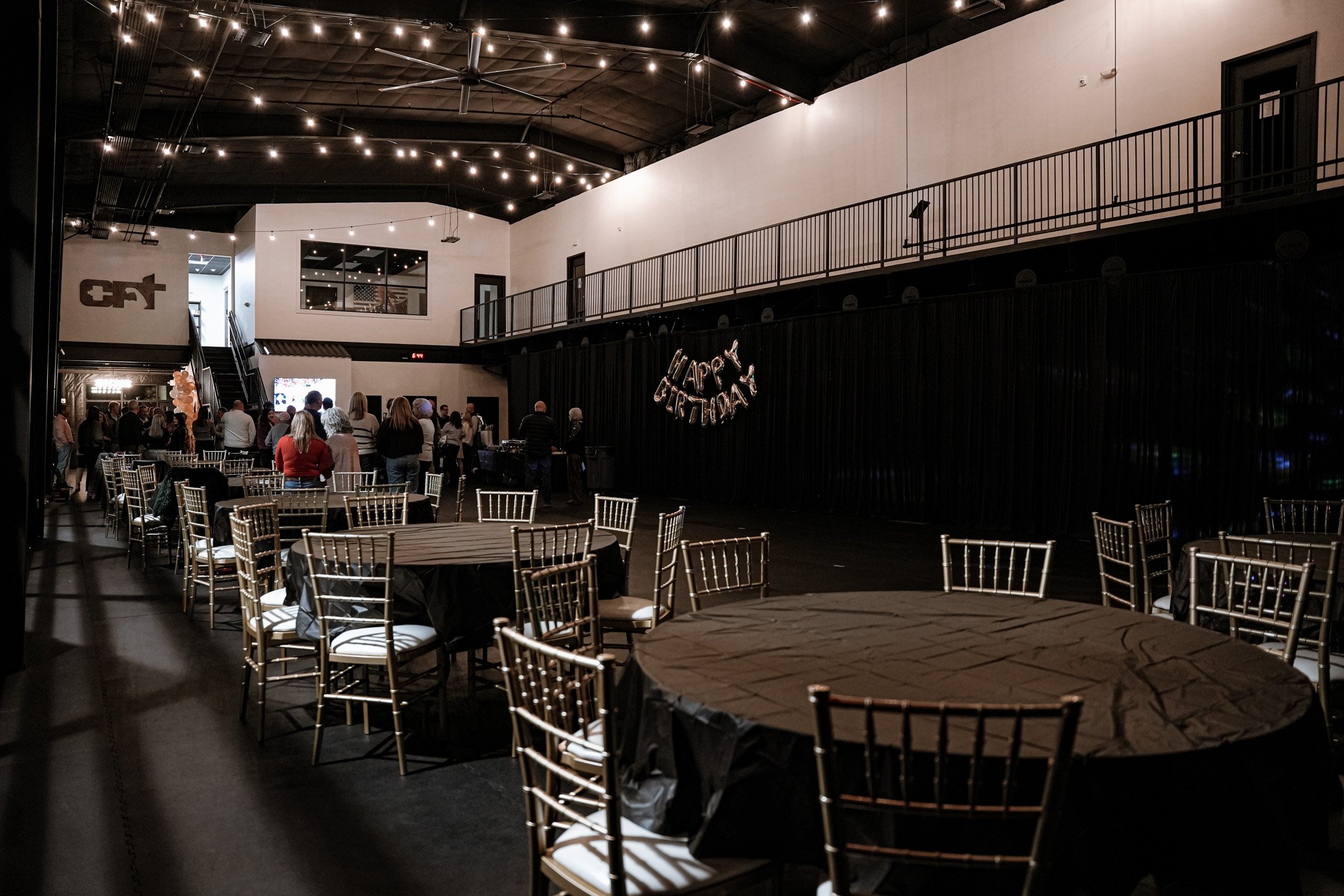 People gathered in a dimly lit event space with round tables and gold chairs, decorated with a "Happy Birthday" balloon banner on the dark wall.