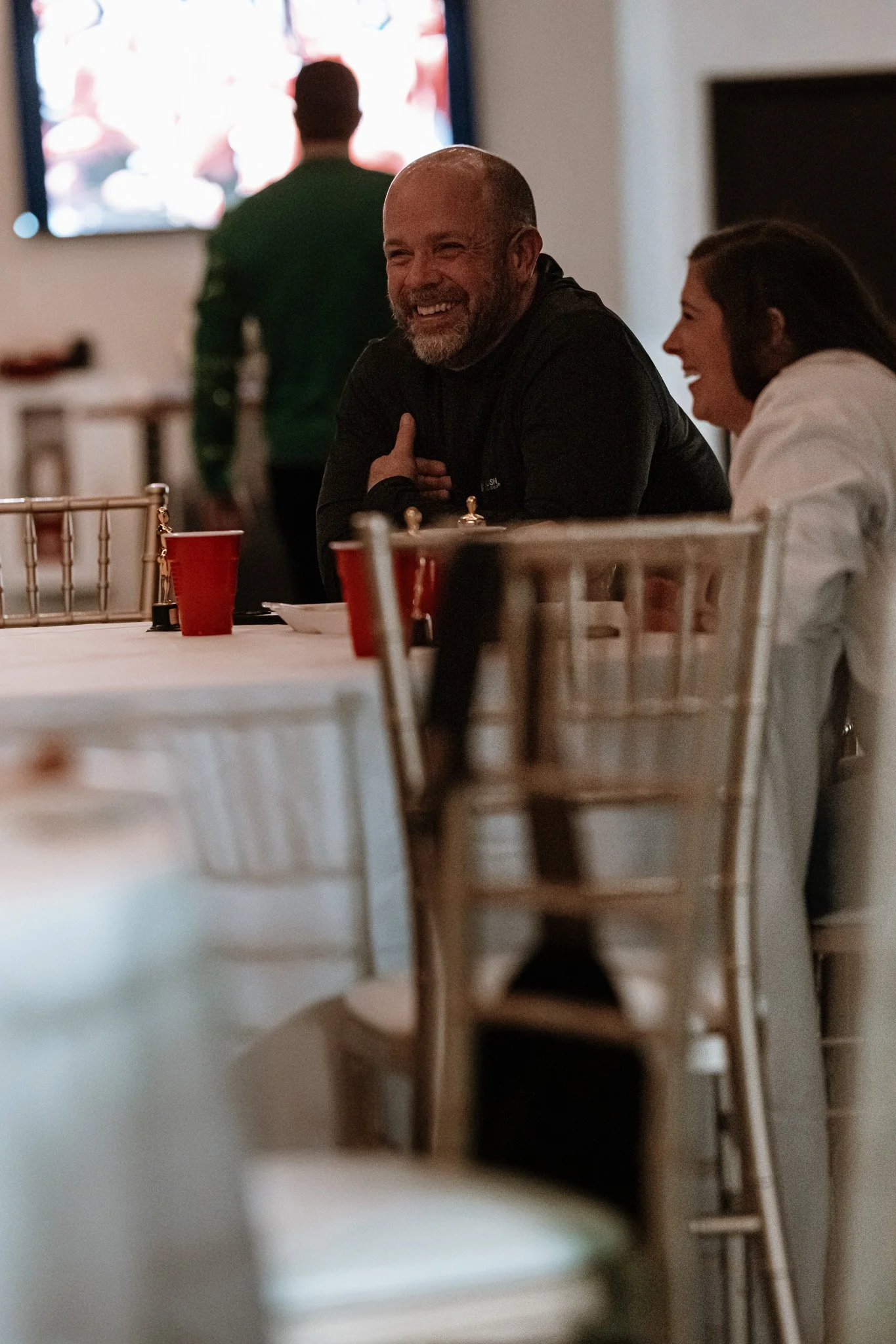 People sitting at a table in a dimly lit room, smiling and enjoying conversation, with a television screen in the background.