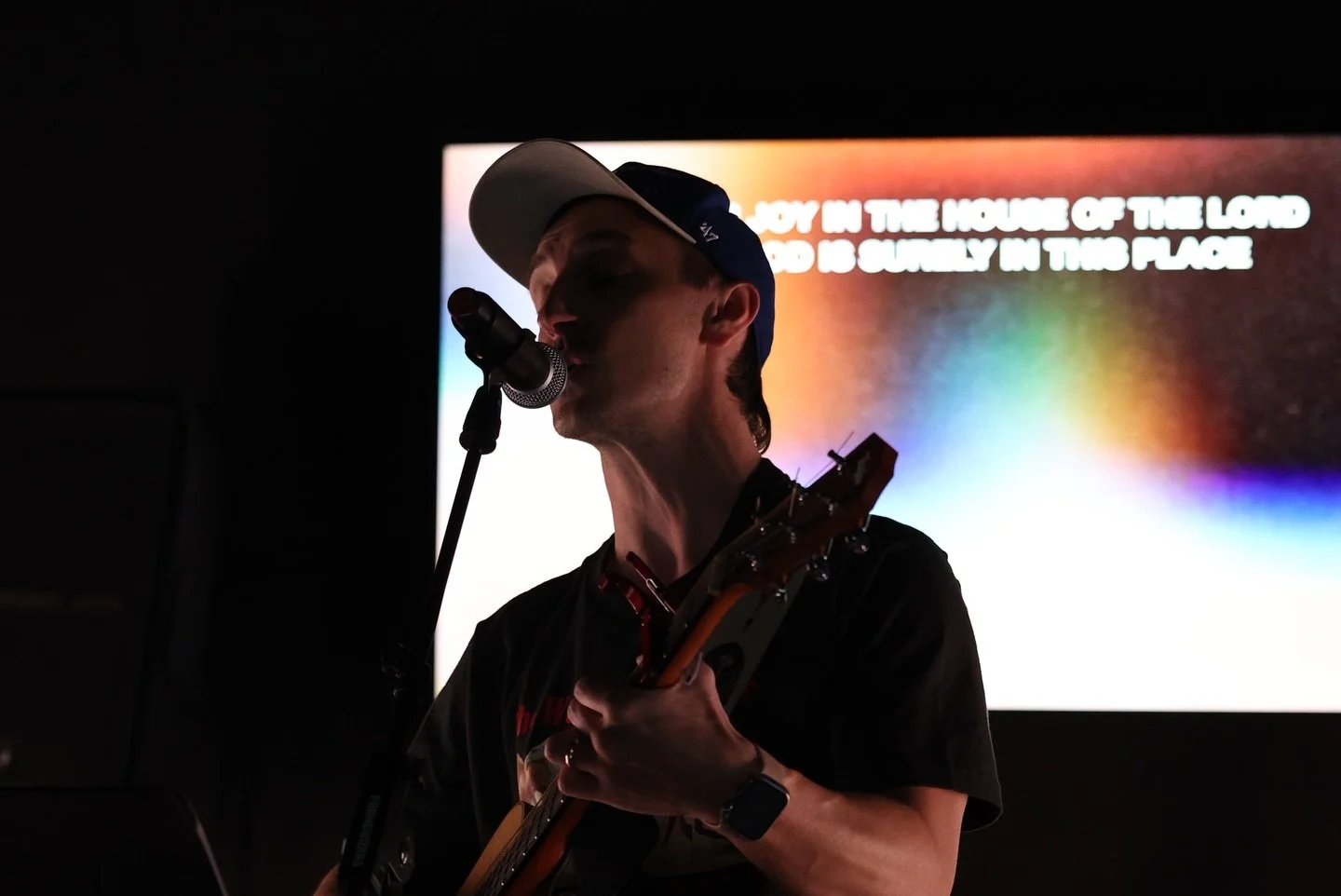 A person singing into a microphone while playing a guitar in a dark room, with a colorful screen in the background.