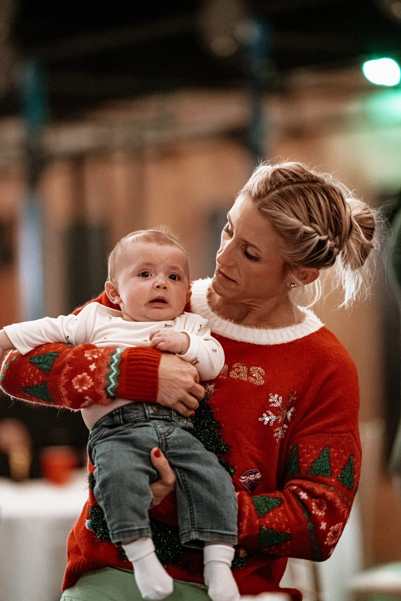 A woman holding a surprised-looking baby indoors, both wearing festive Christmas sweaters with holiday patterns.