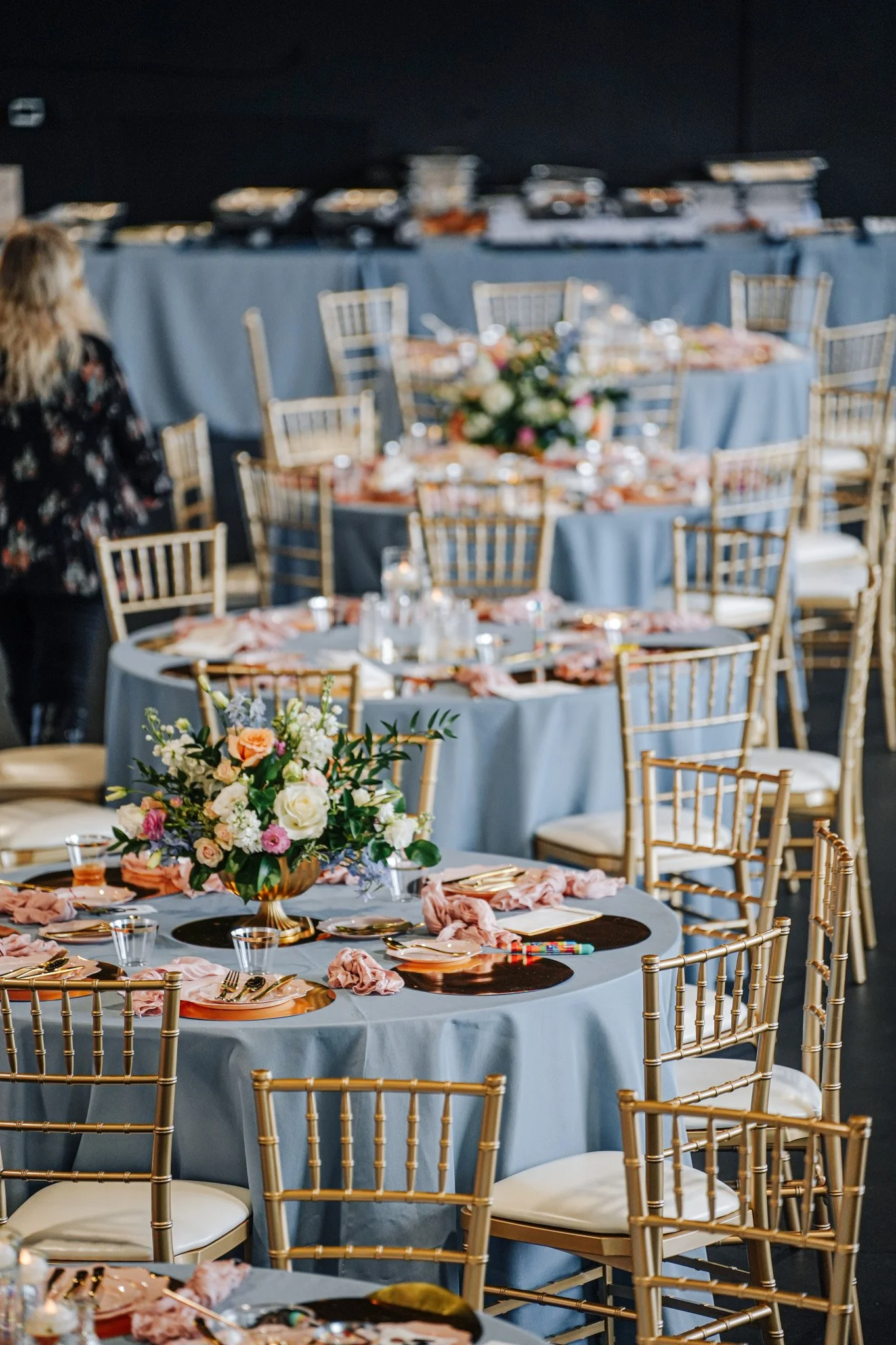 Elegant banquet table with a centerpiece of flowers, set with plates, glasses, and cutlery, with gold chairs around various tables in a decorated event space.