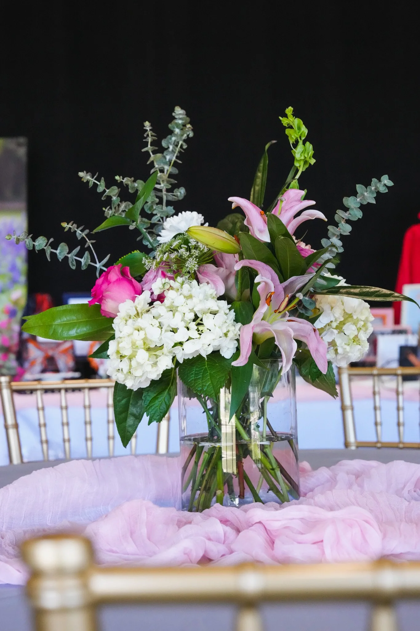 A flower arrangement in a clear glass vase on a table with pink cloth, containing pink lilies, white hydrangeas, and various green foliage, against a black background.