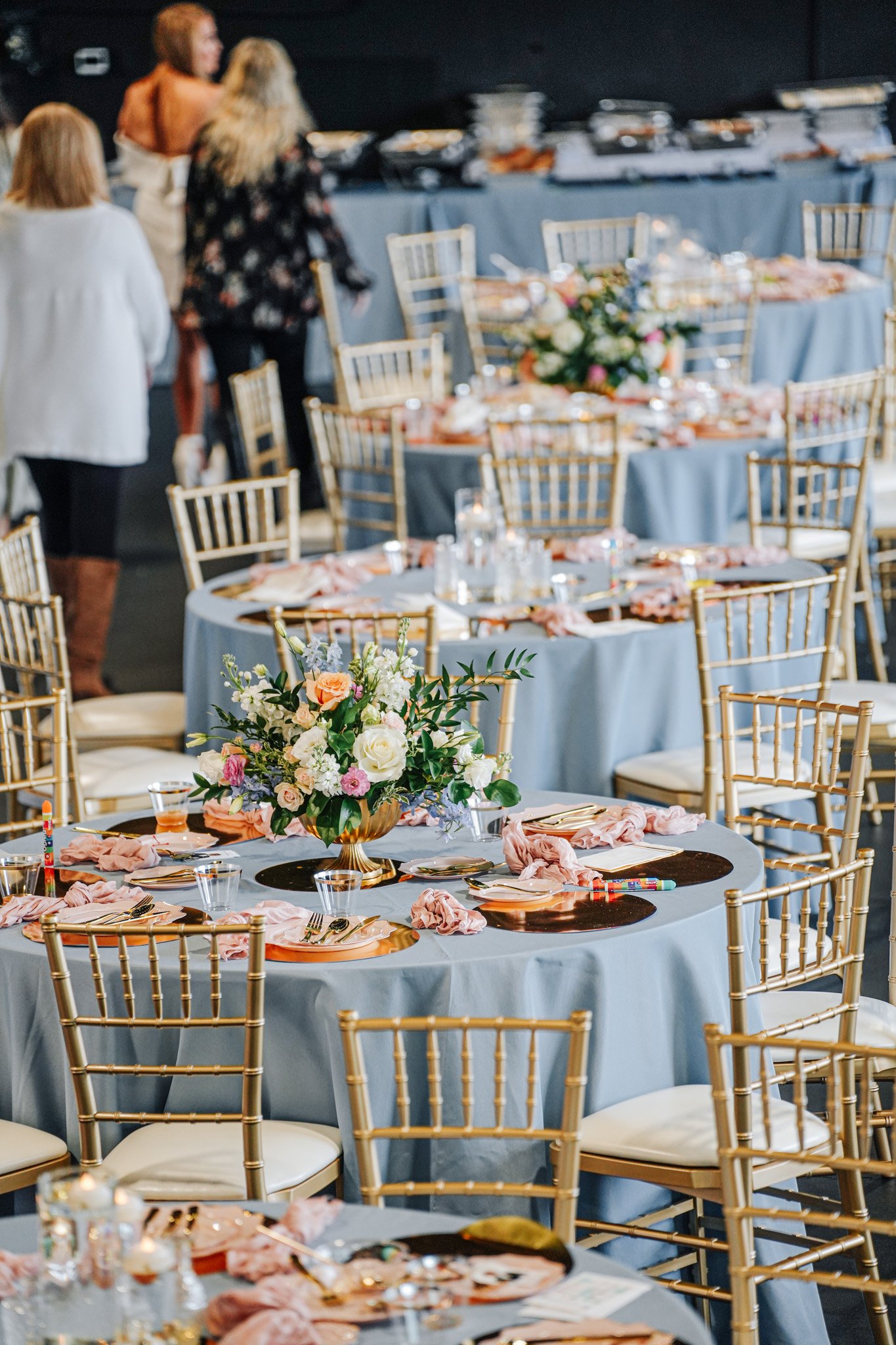 Empty banquet tables decorated with pink cloth napkins, floral centerpieces, and gold chairs, set up for a wedding or event.