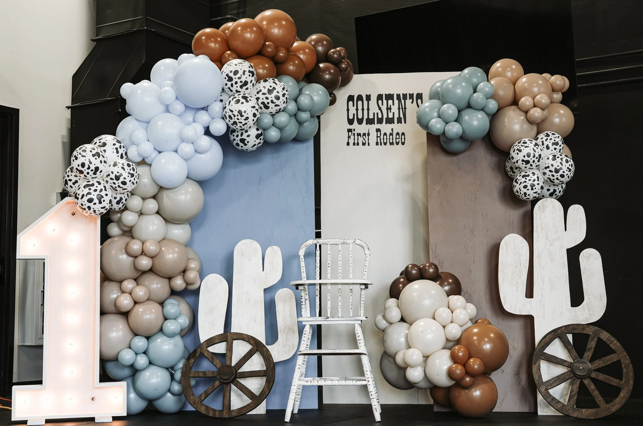 Decorative backdrop with balloons, wooden cacti, a white chair, and a large illuminated number one, at Colsen's First Rodeo event.